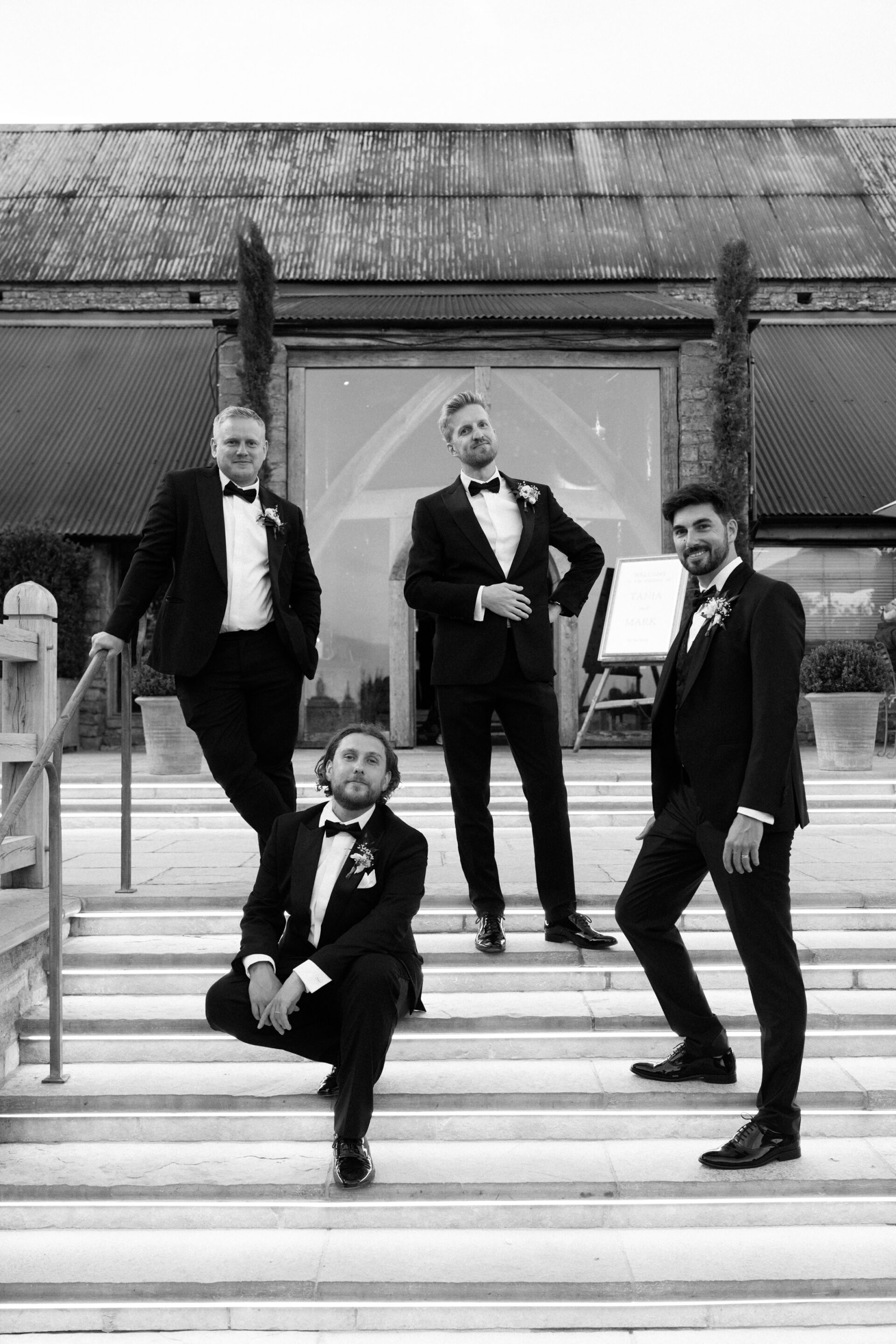 Four men in tuxedos are on outdoor steps by a modern barn-like building; three are standing and one is sitting.