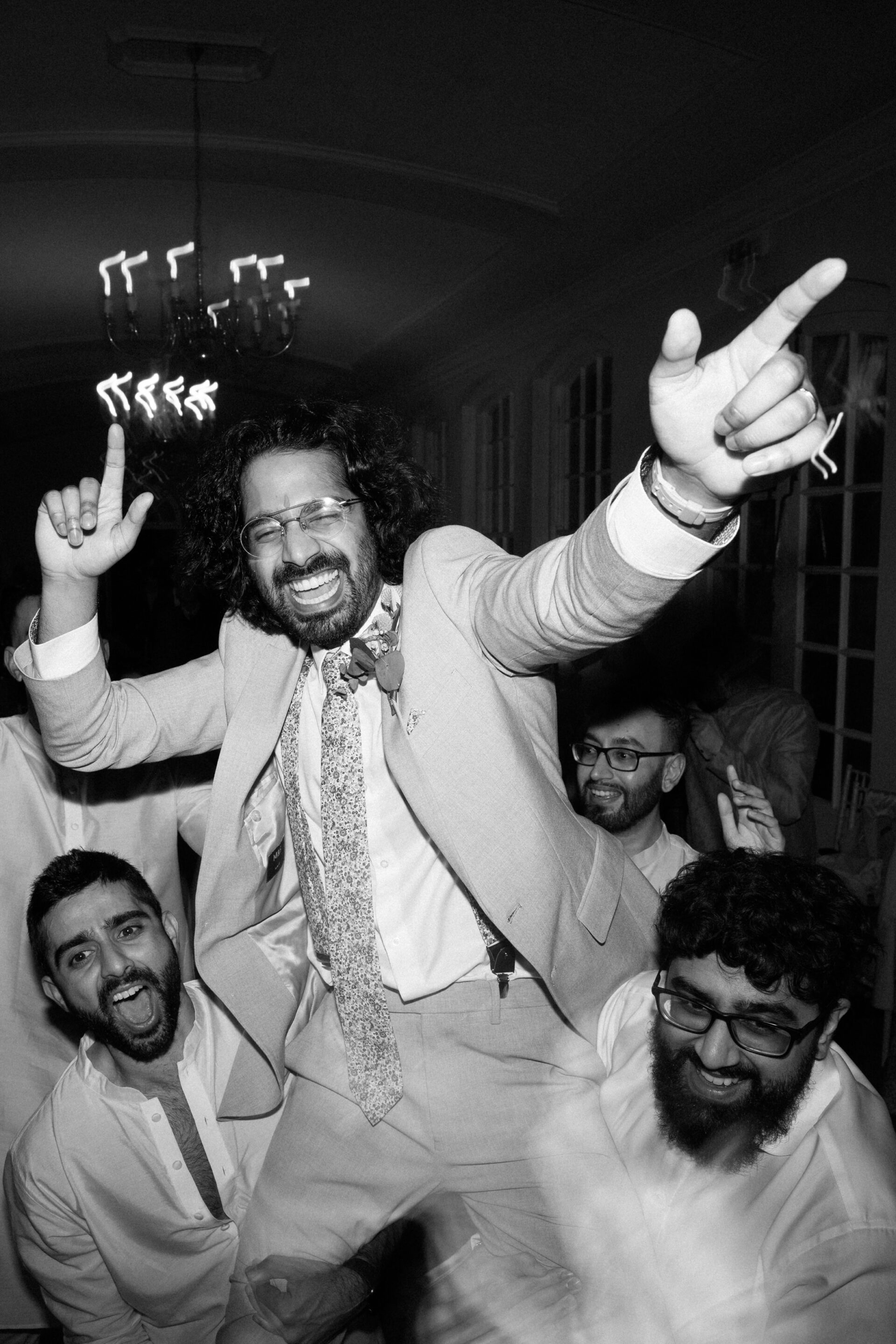 Friends are lifting a man in a suit at a party, everyone looks happy and excited with their hands up inside a busy room.