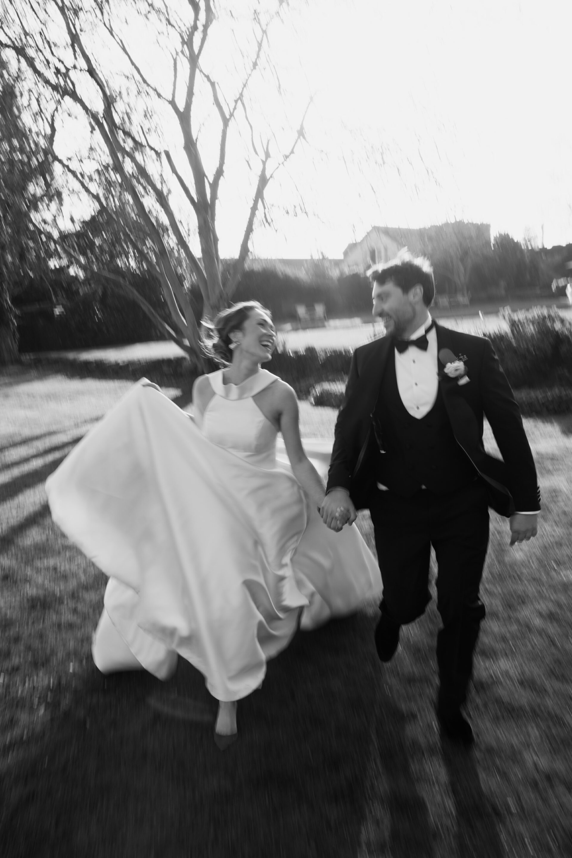 A bride in a white dress and a groom in a tuxedo run hand in hand outside, both smiling as they run with motion blur around them.