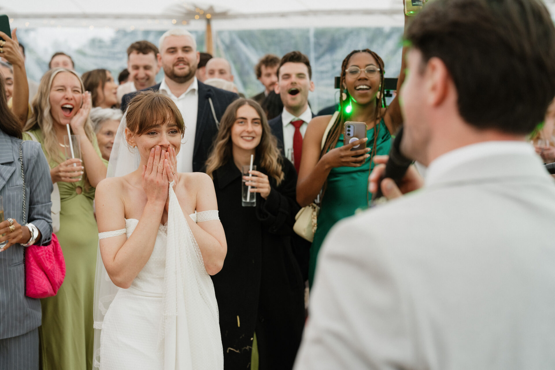 A bride stands surprised with her hand over her mouth as a man in a suit faces her, and guests smile and clap at the wedding.