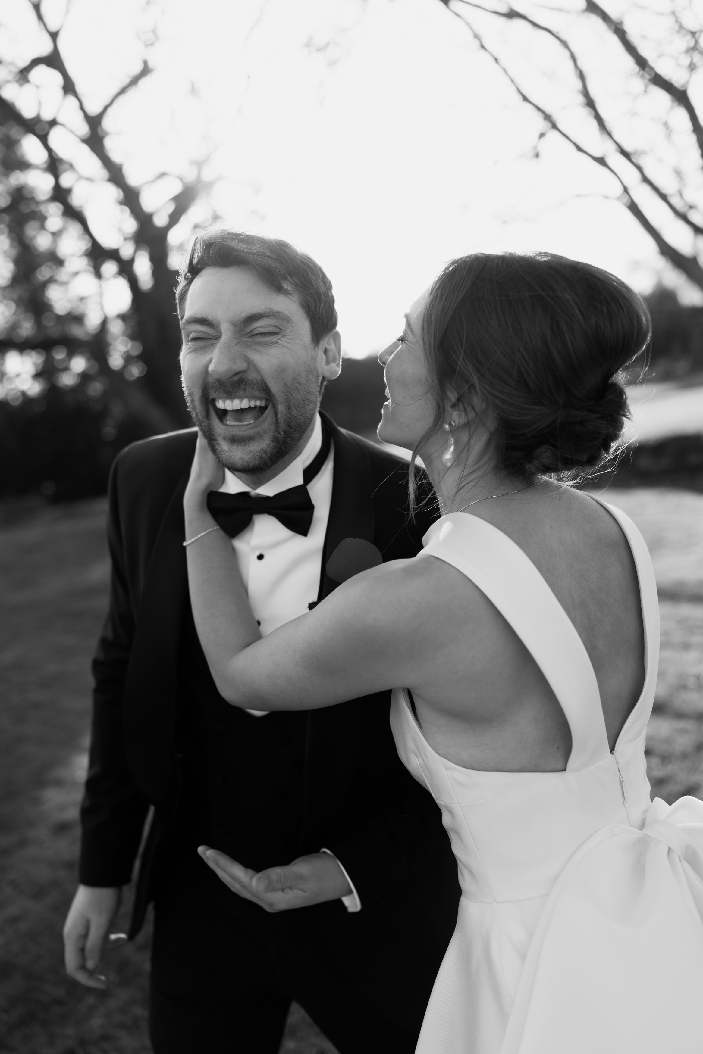 A bride in a white dress gently holds her groom’s face as he laughs, both outside with trees and sunlight behind them.