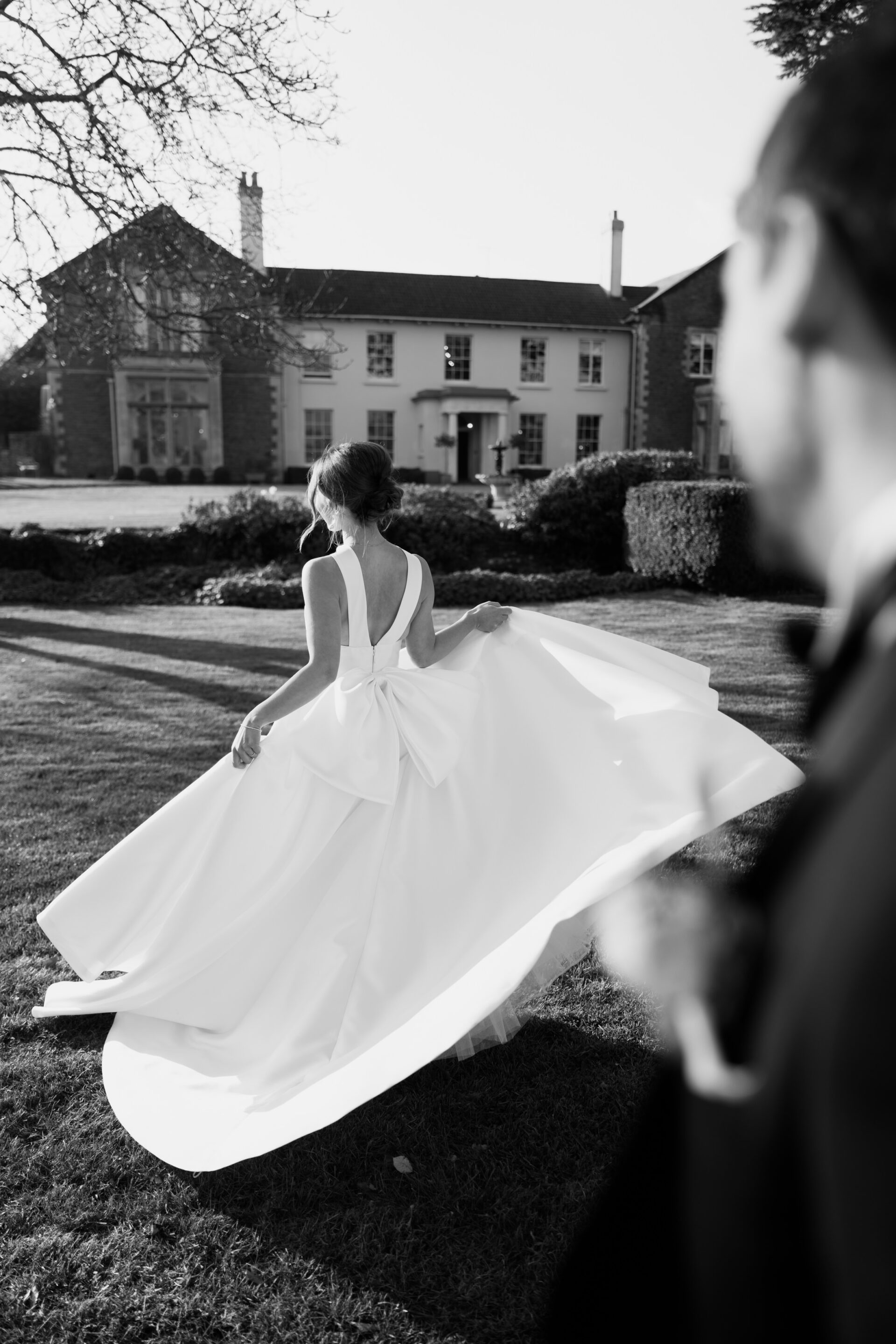 A woman in a white wedding dress walks on the grass outside a large house, with a man in a suit nearer to the camera. It’s in black and white.