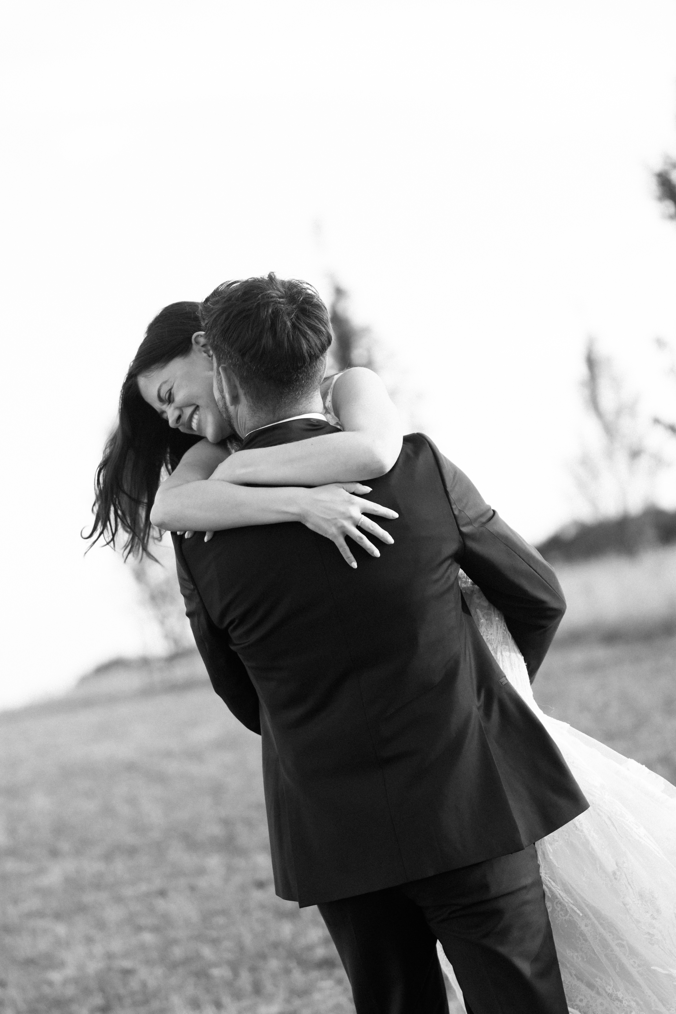 A man in a suit picks up and hugs a woman in a wedding dress outside. They're smiling, with grass and trees behind them.
