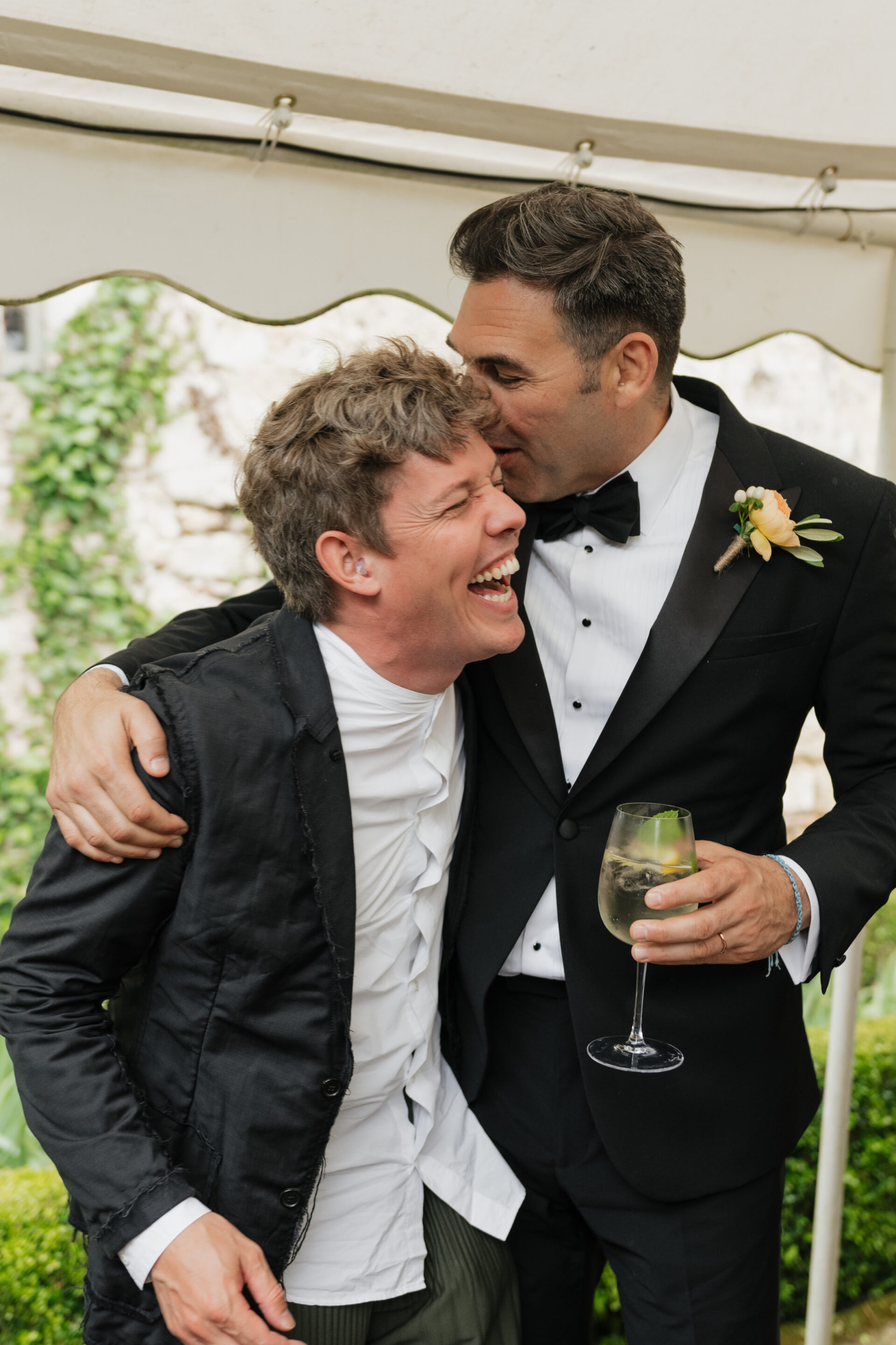 Two men in suits enjoy a happy moment; one is laughing while the other hugs him and holds a glass of white wine.