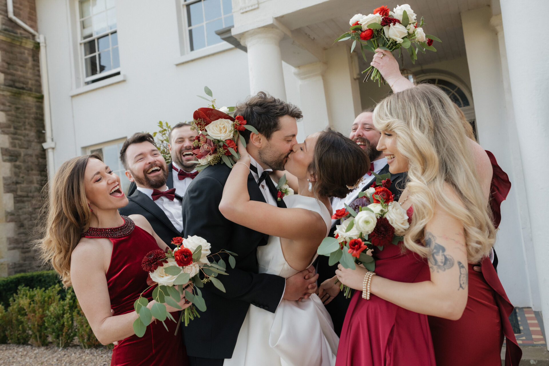 A bride and groom share a kiss outside while their happy wedding party in red dresses and suits holds red and white flower bouquets.