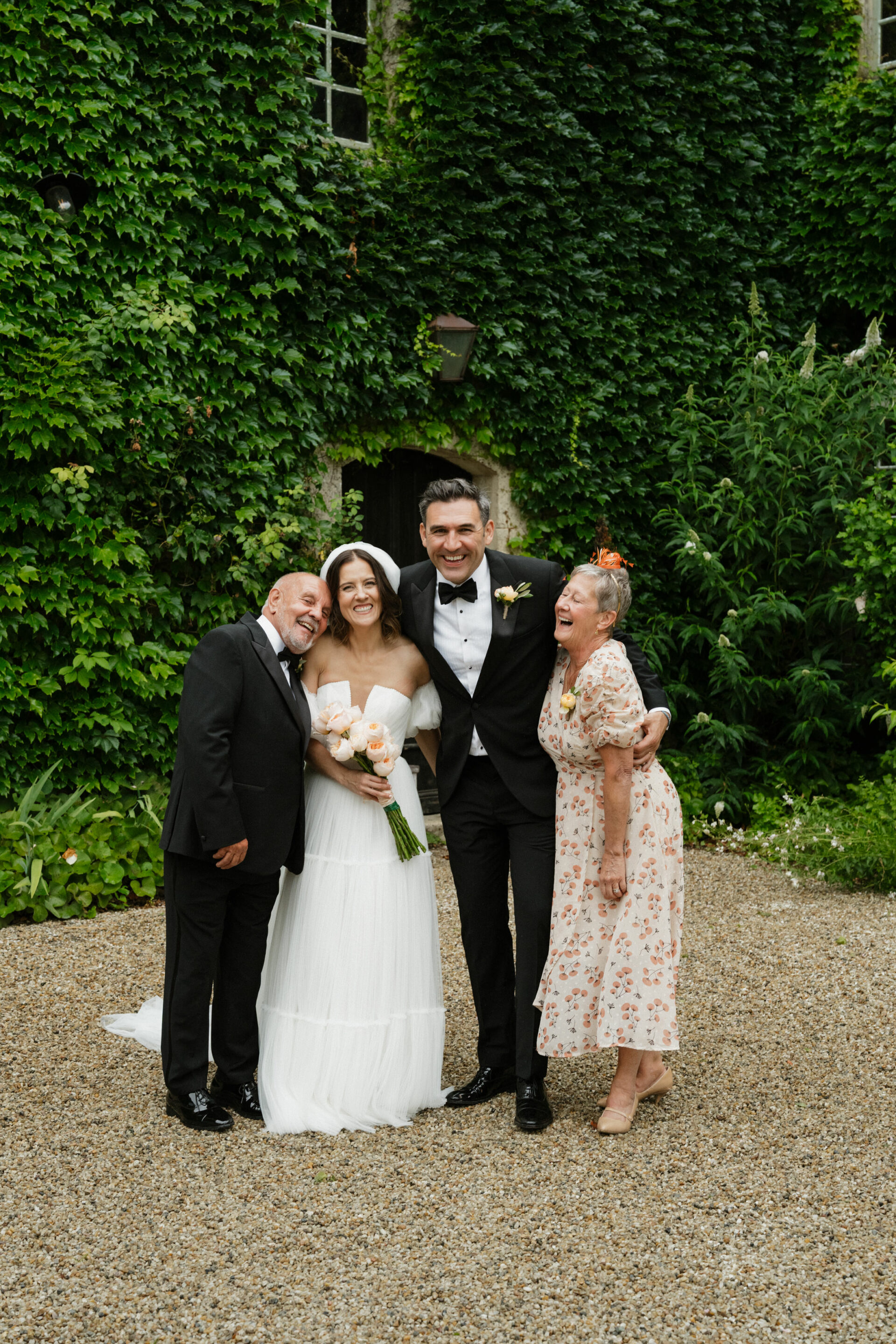 Four people are standing outside by a wall covered in green ivy. A bride and groom are in the middle, with parents on each side.