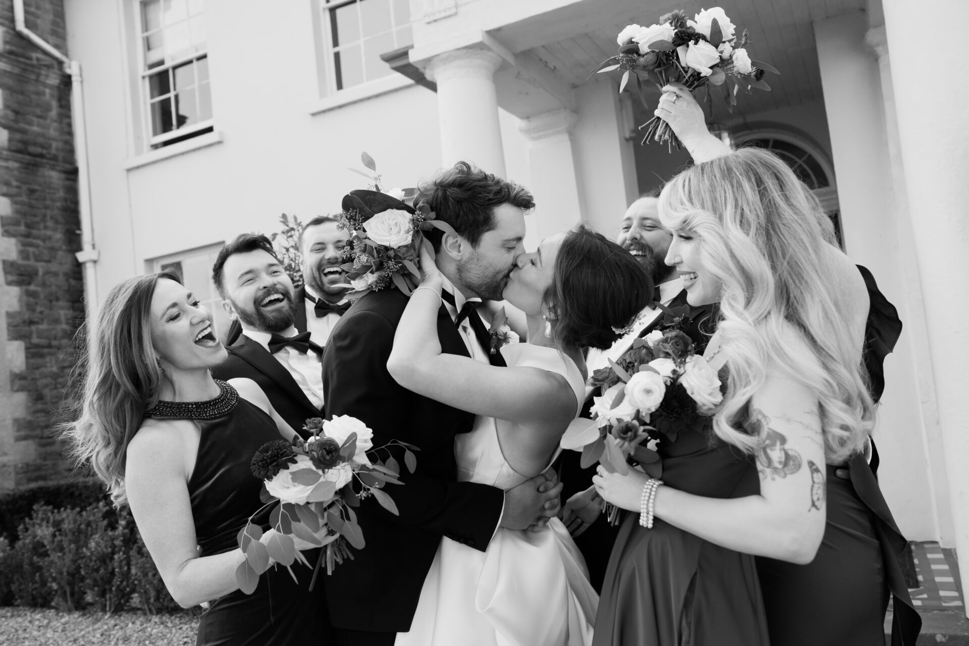 A bride and groom share a kiss outside while their happy, well-dressed friends stand around them holding flowers.