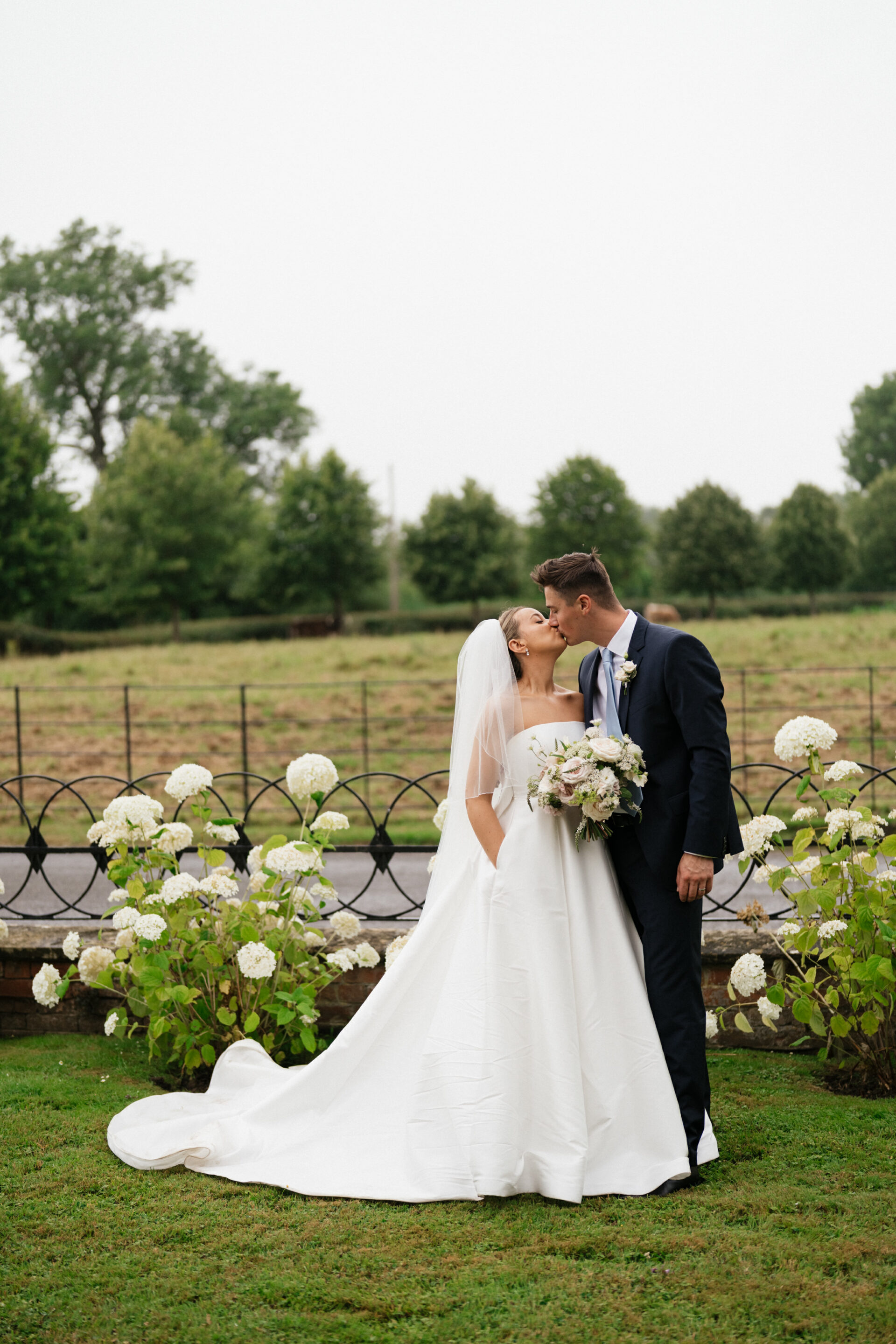 A bride in a white dress and veil kisses her groom, who wears a dark suit and holds flowers. They're outside by white blooms.