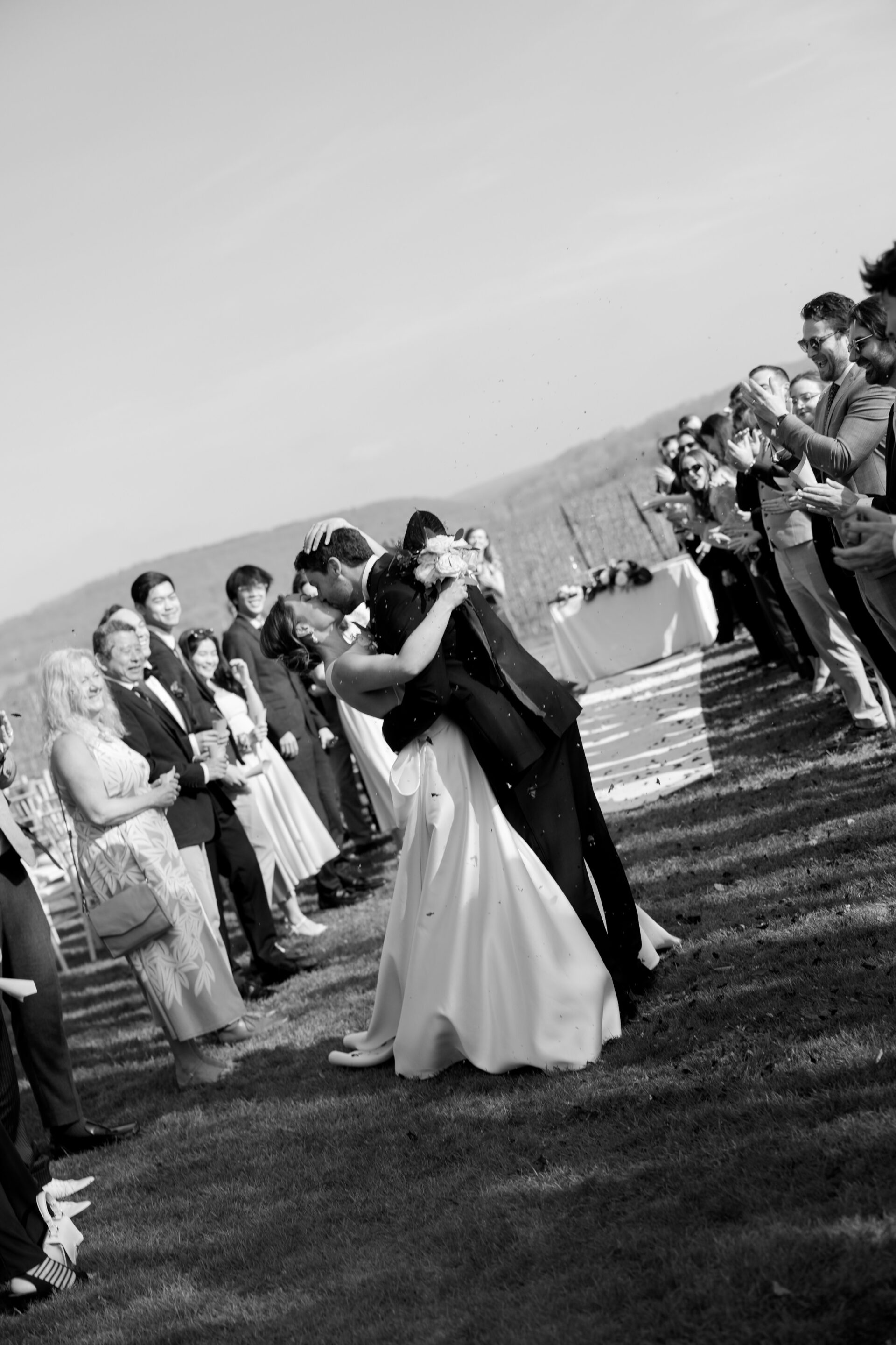 The bride and groom share a kiss outside at their wedding as guests on both sides clap and watch.