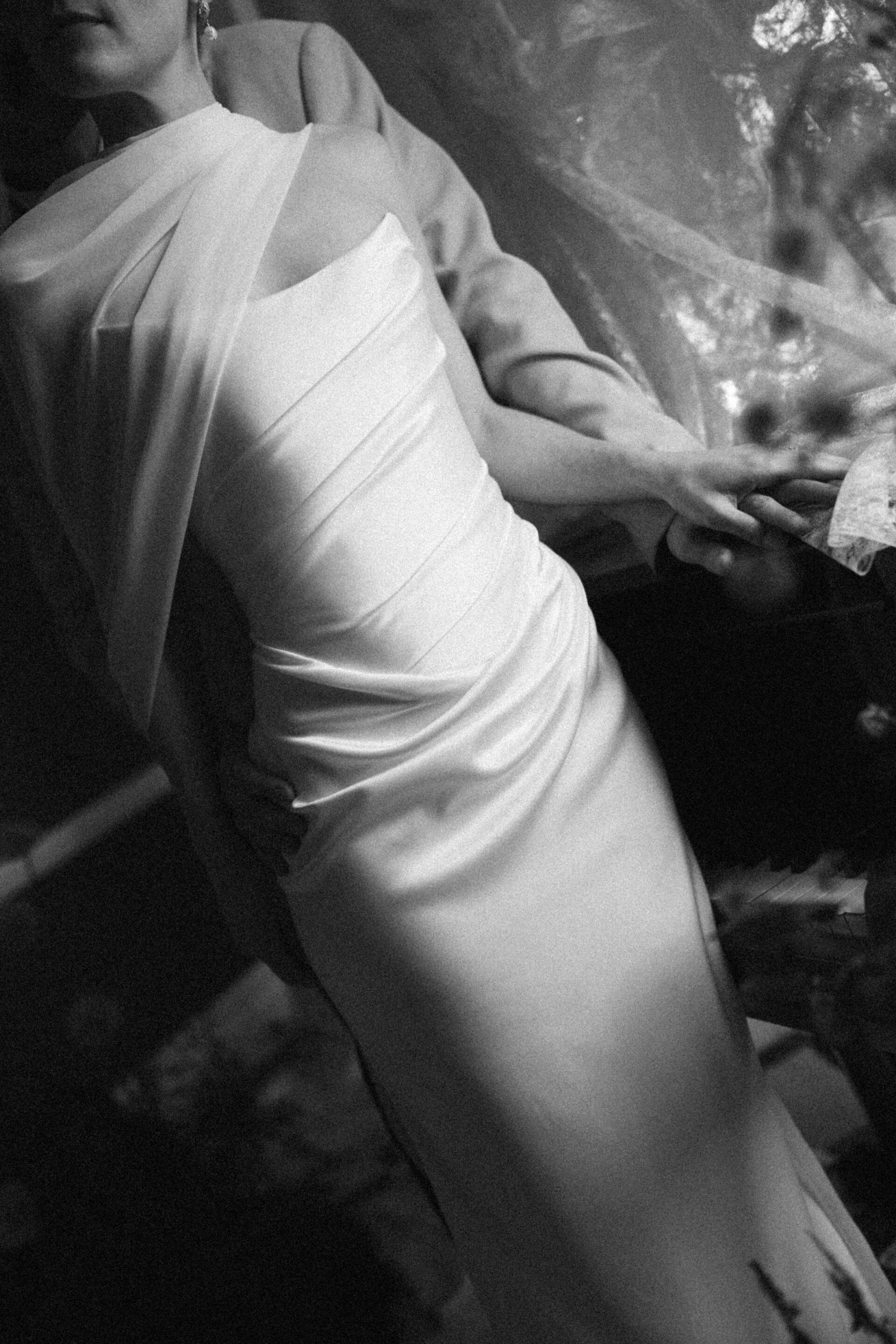 Someone in a white, draped dress stands while another person behind them rests their arm across the front, in low lighting.