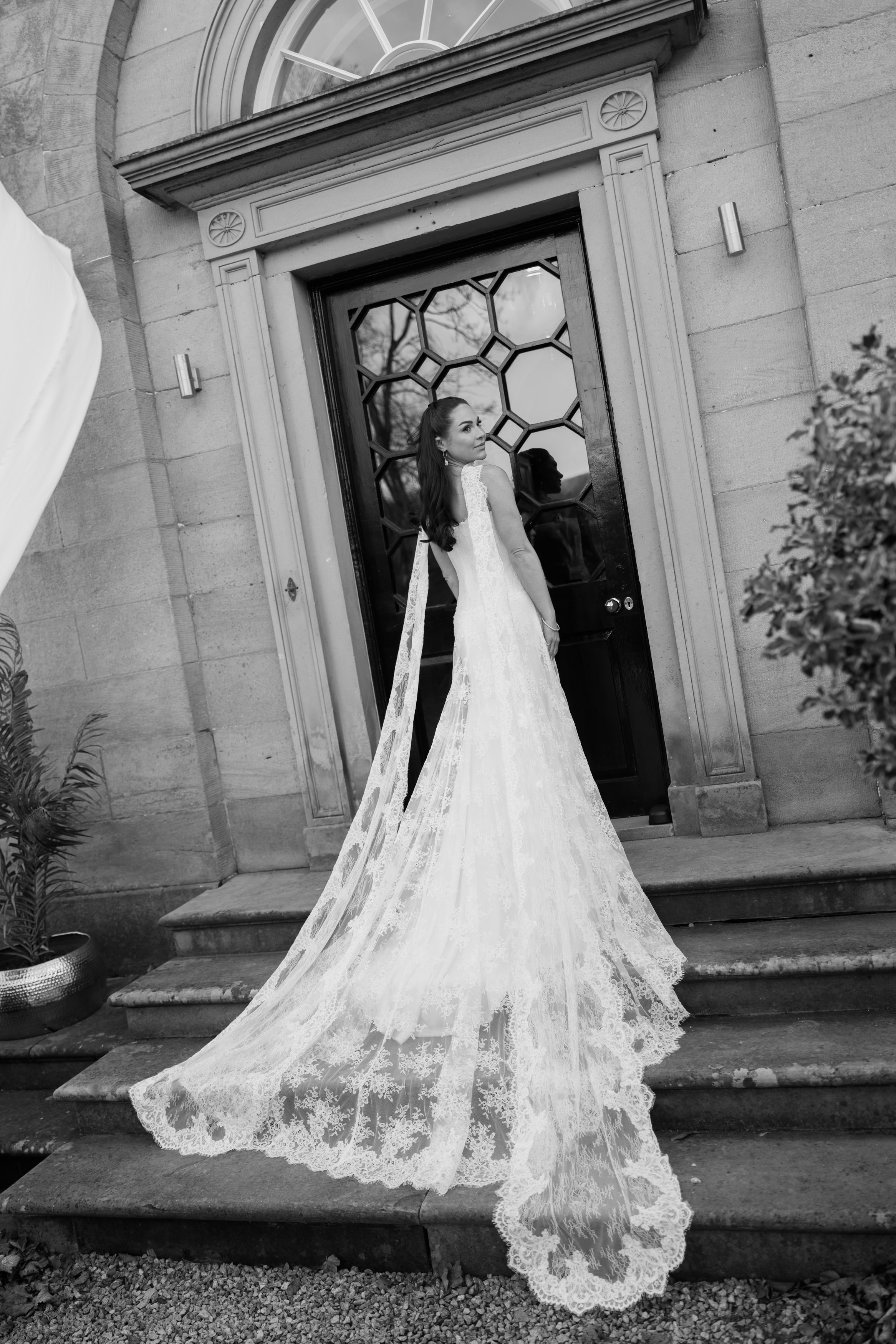 A woman wearing a long lace wedding dress stands on stone steps by a big door, glancing back over her shoulder.