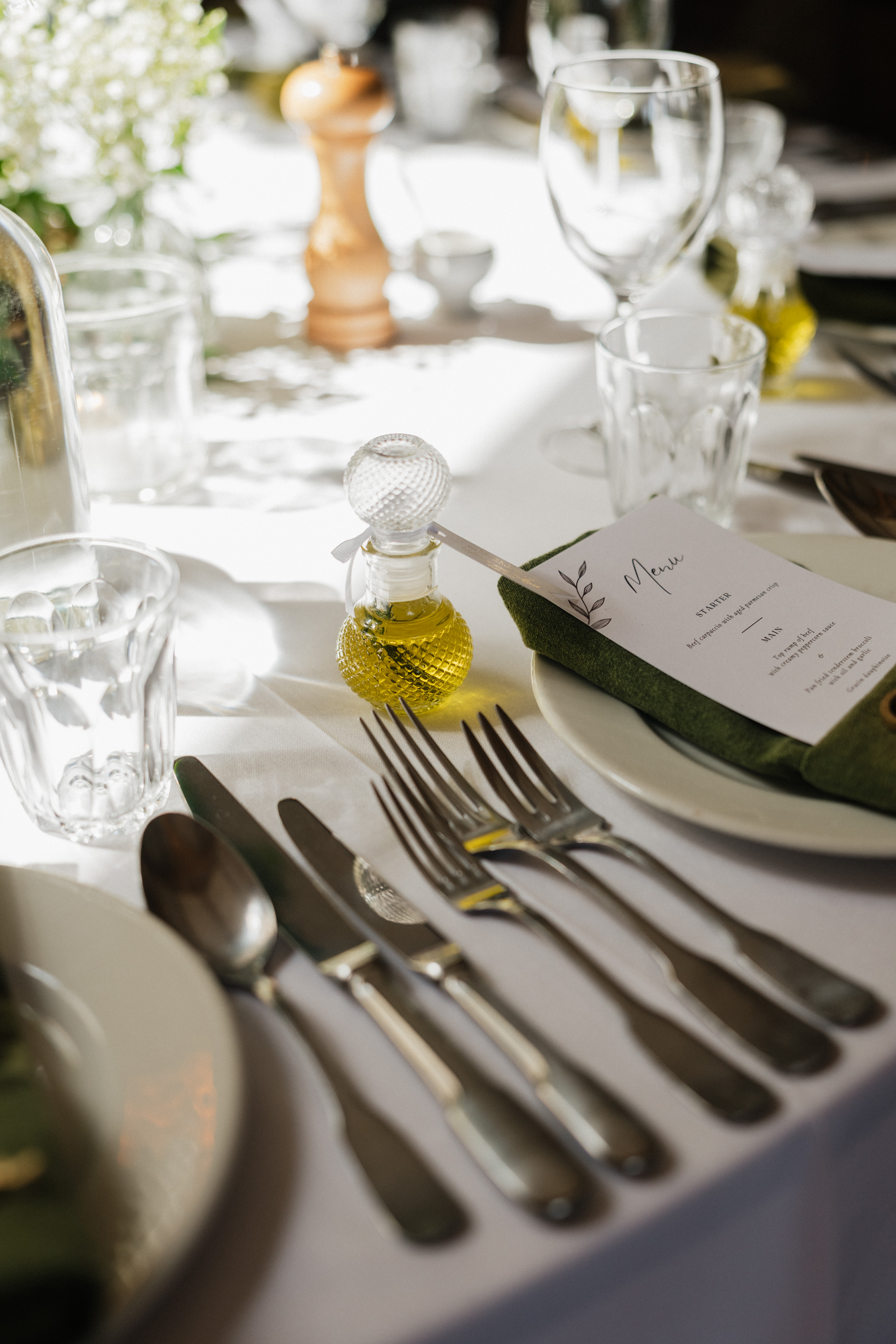 A table set with dishes, silverware, a glass, a menu on a green napkin, and olive oil on a white cloth in daylight.