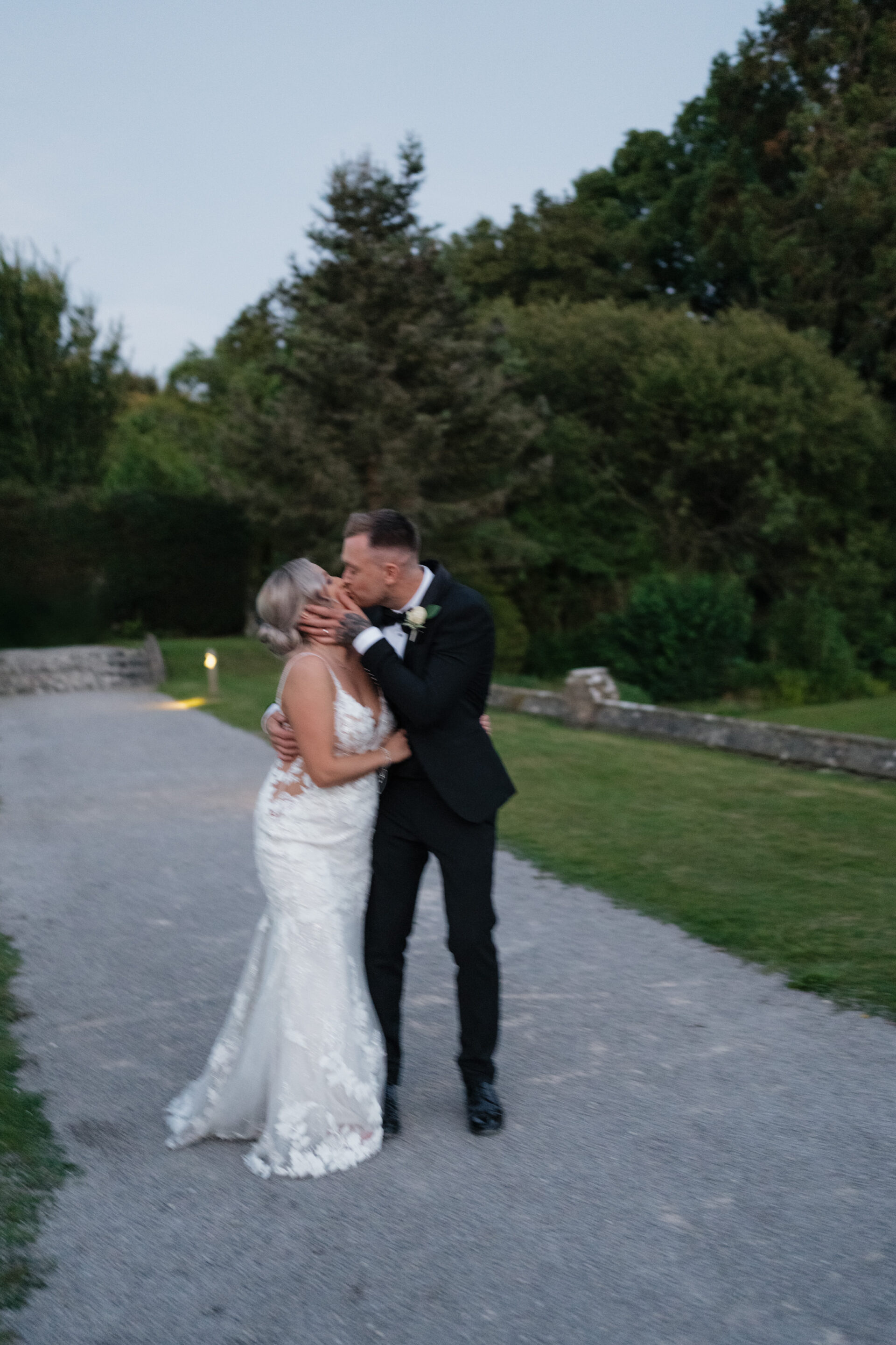 A bride in a white dress and a groom in a black suit kiss outside on a path, with lots of green plants around them.