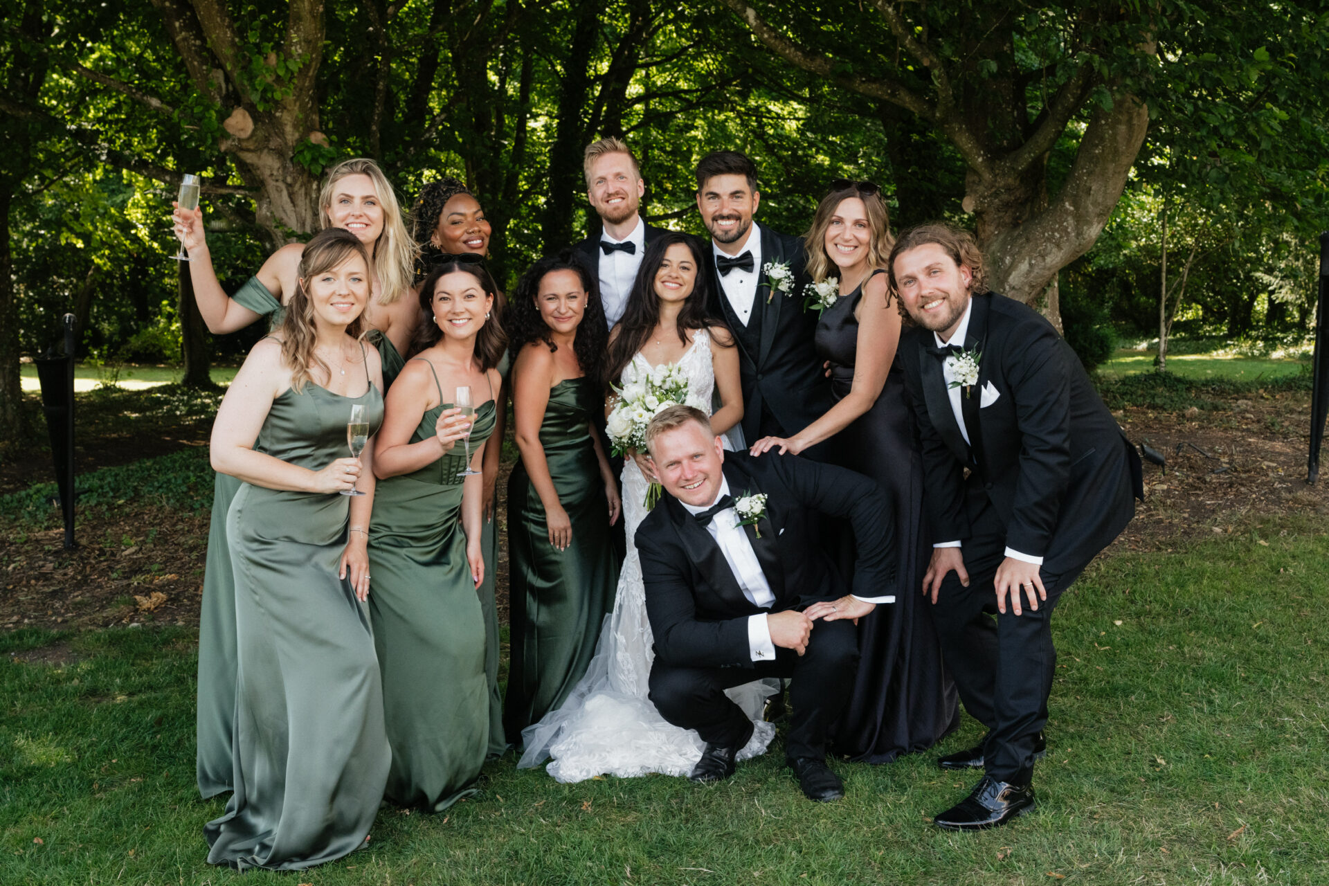 The bride and groom stand outside with their wedding party, as bridesmaids in green dresses and groomsmen in black suits smile for a photo.