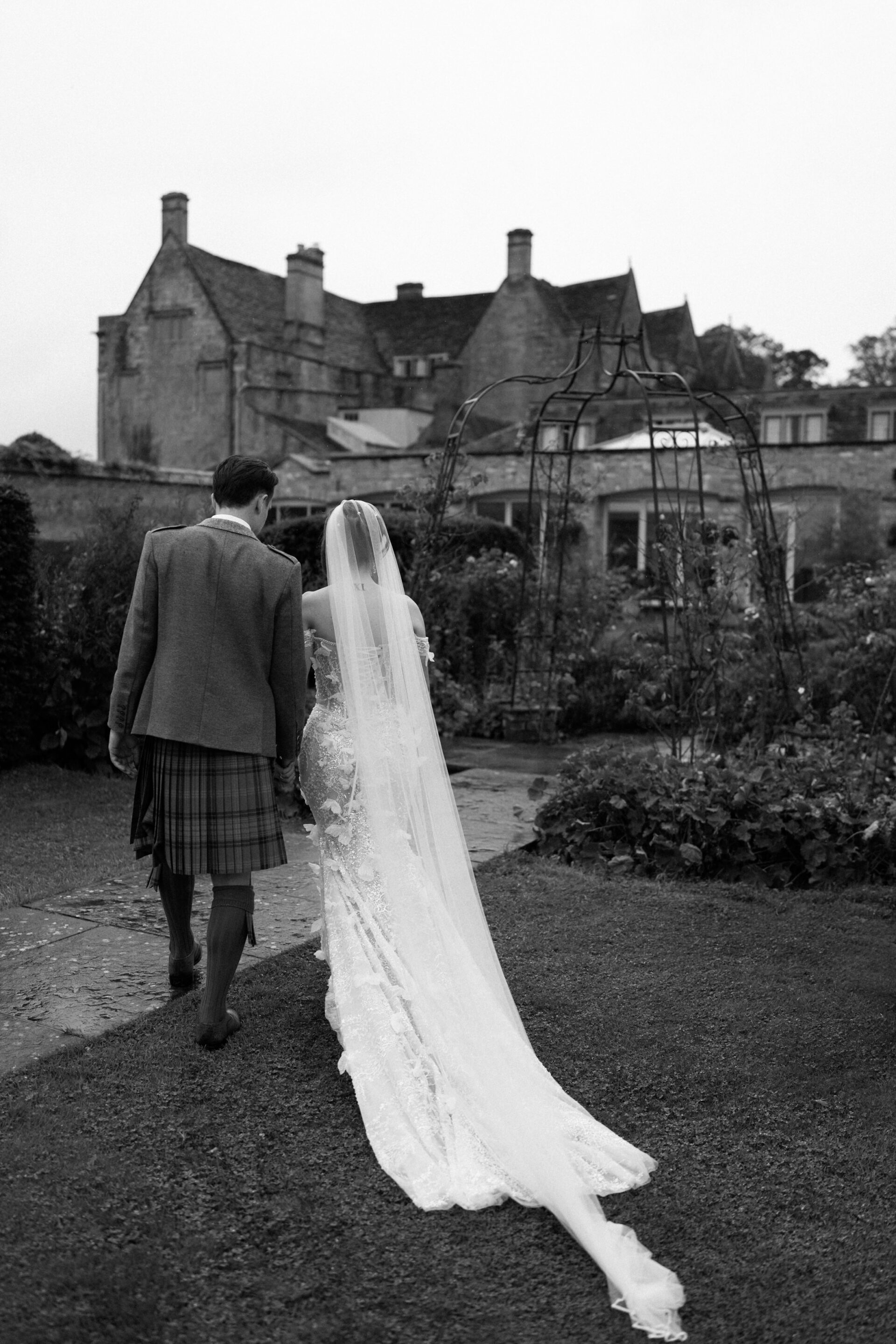 A bride wearing a long veil and a groom in a kilt walk through a garden, with a big stone building behind them.