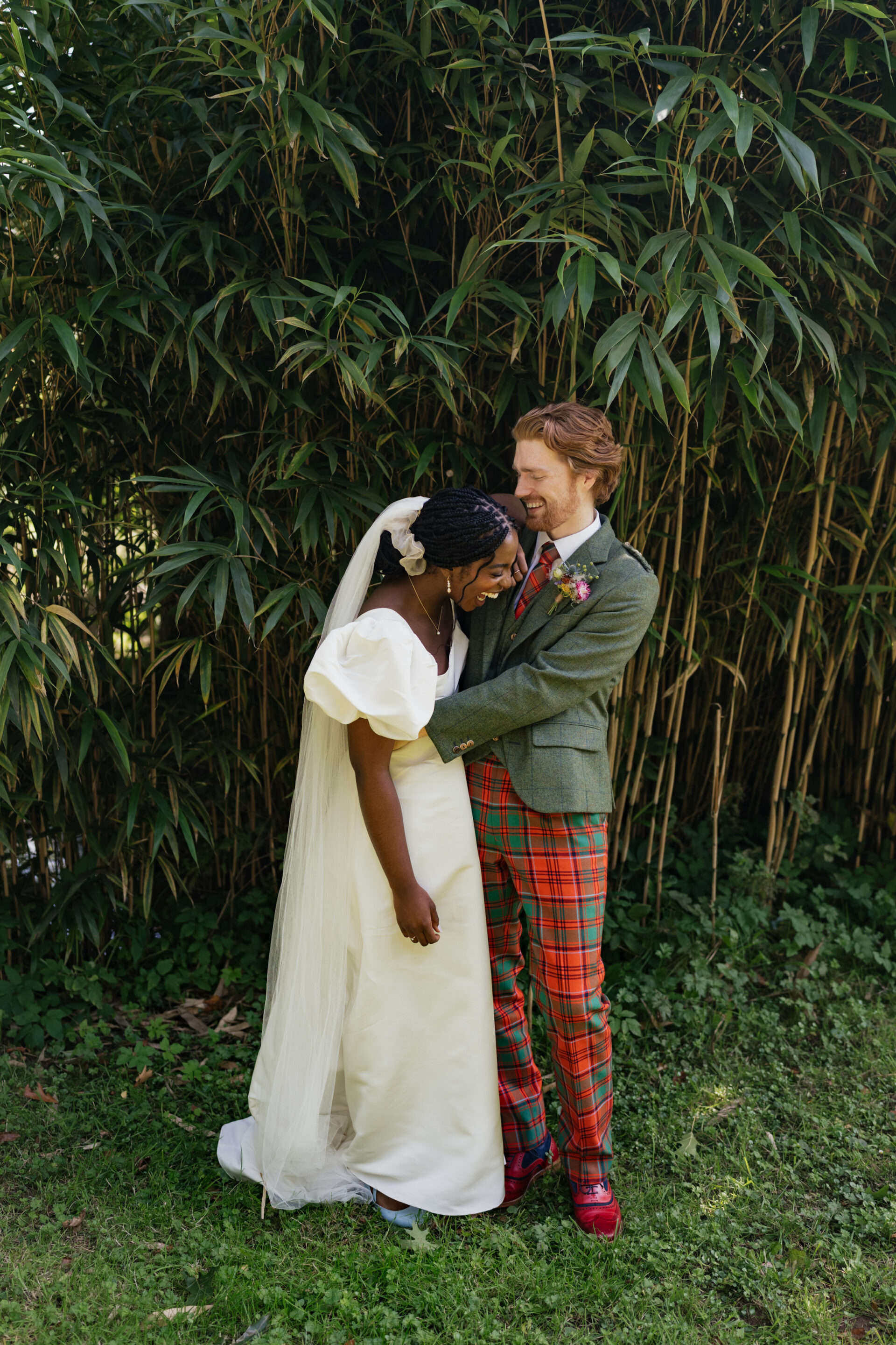A bride in a white dress and a groom wearing a green jacket and red plaid pants smile together by some tall green bamboo plants.