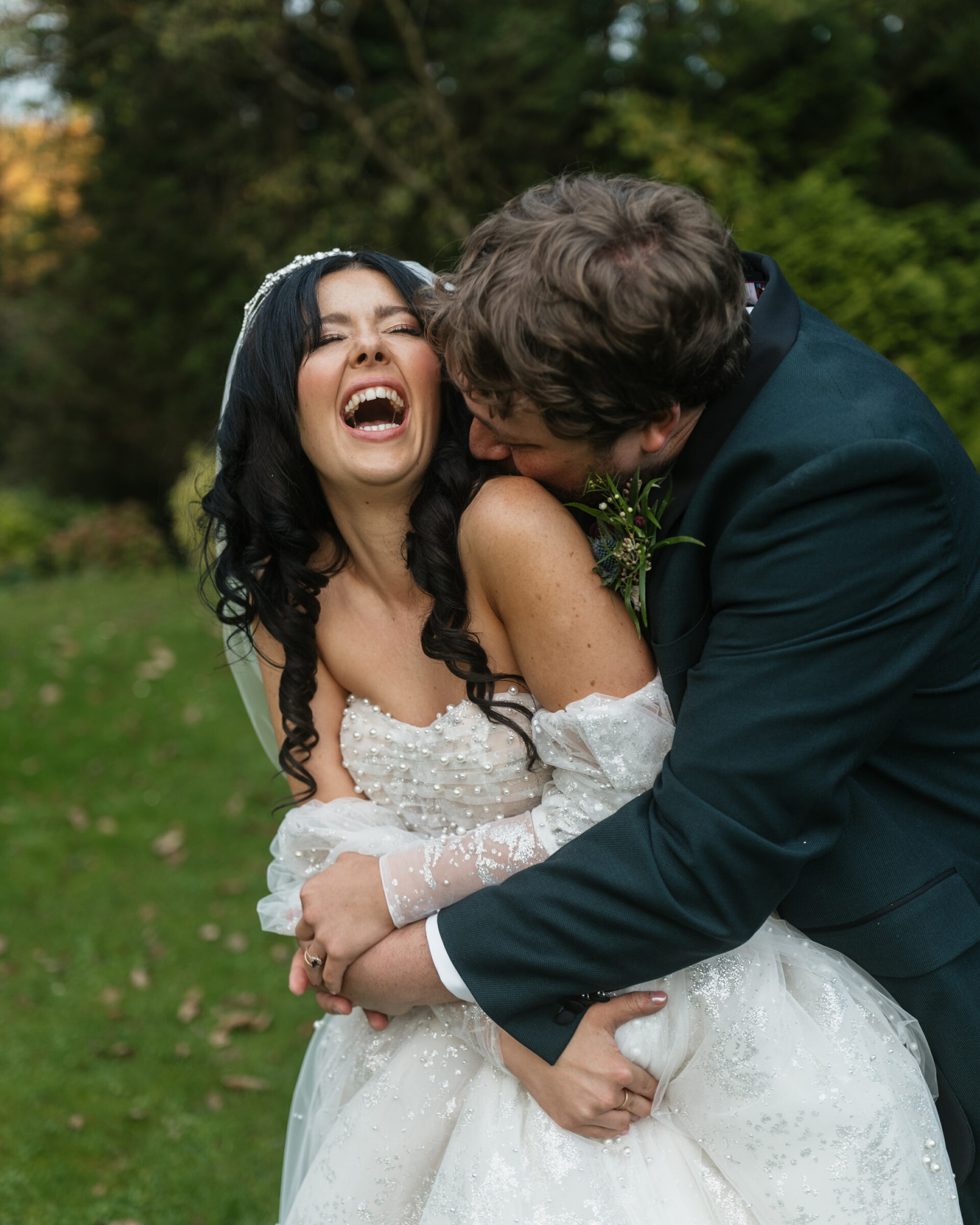 A bride in a white dress and a groom in a dark suit hug and laugh together on the grass outside, with trees behind them.