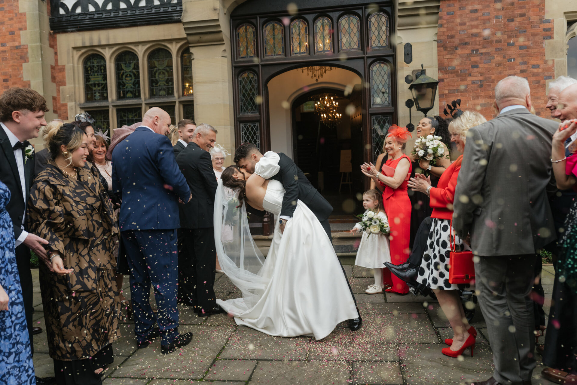 A bride and groom kiss outside a building as their guests, dressed up, toss confetti and cheer around them.
