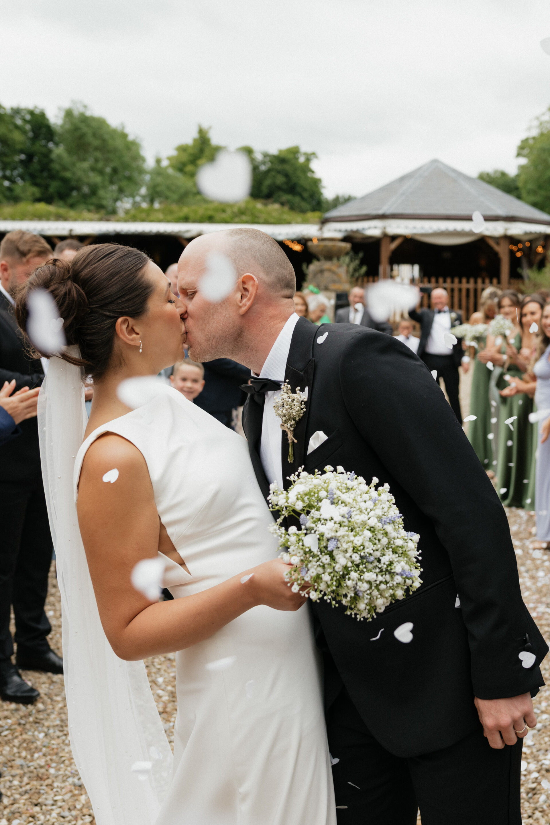 A bride and groom share a kiss outside, with guests around them and flower petals falling after their wedding ceremony.