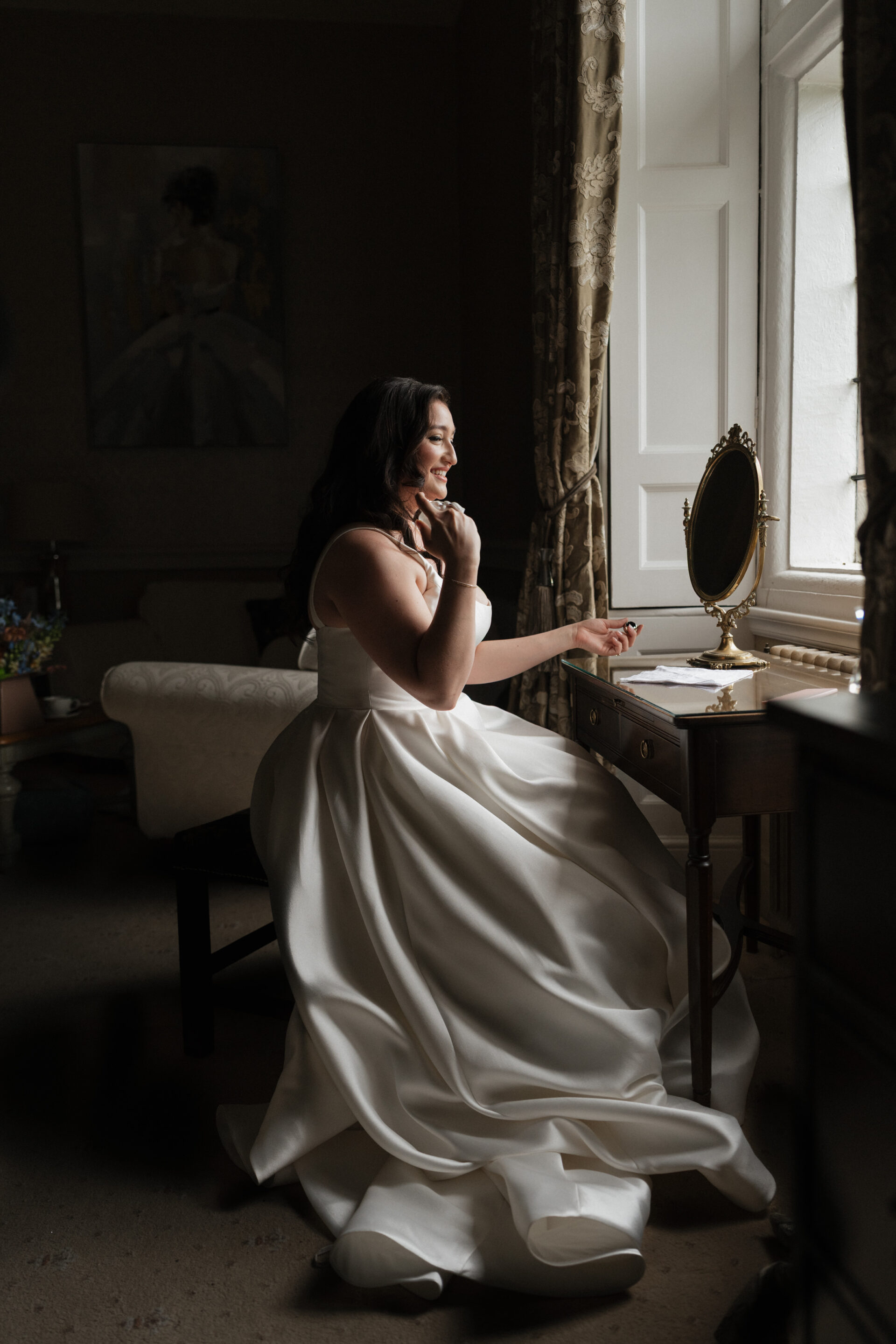 A woman in a white wedding dress sits by a window at a vanity table, smiling as she puts on her earring.