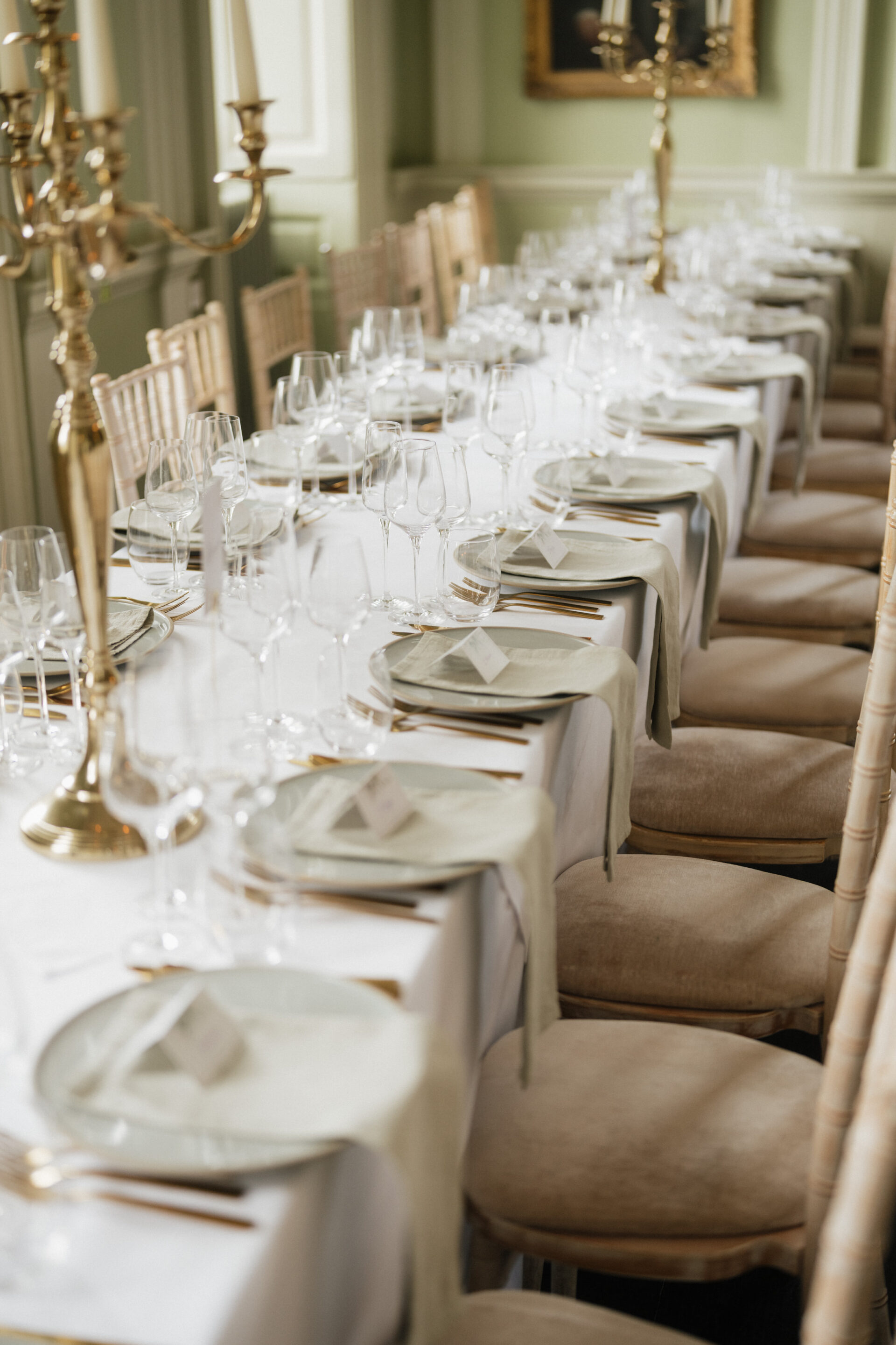 A long dining table with a white tablecloth, dishes, glasses, gold utensils, name cards, and beige chairs in a fancy room with candelabras.