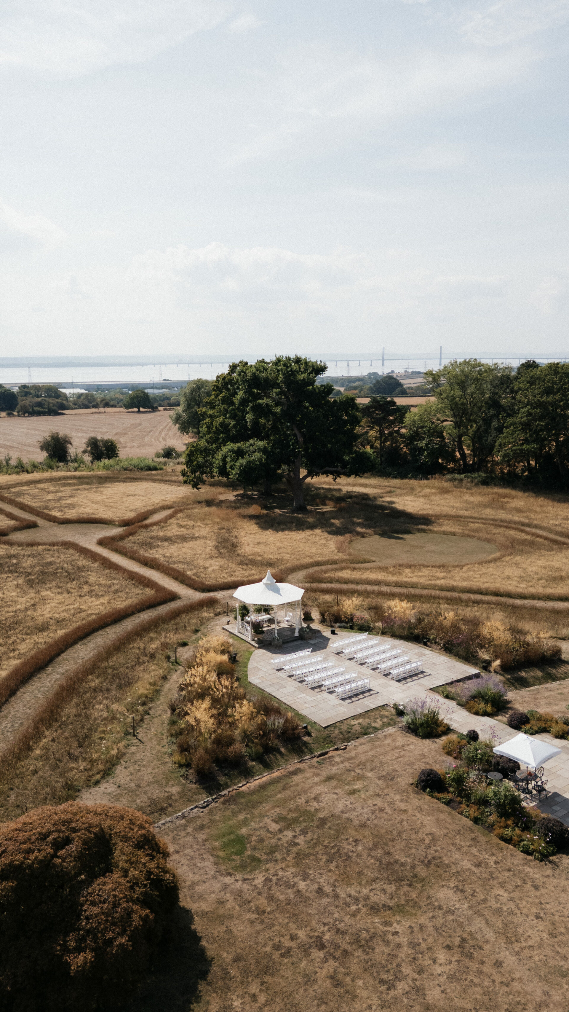 Looking down at an outdoor wedding with white chairs and a canopy, set among dry grass fields, paths, and a bridge in the distance.