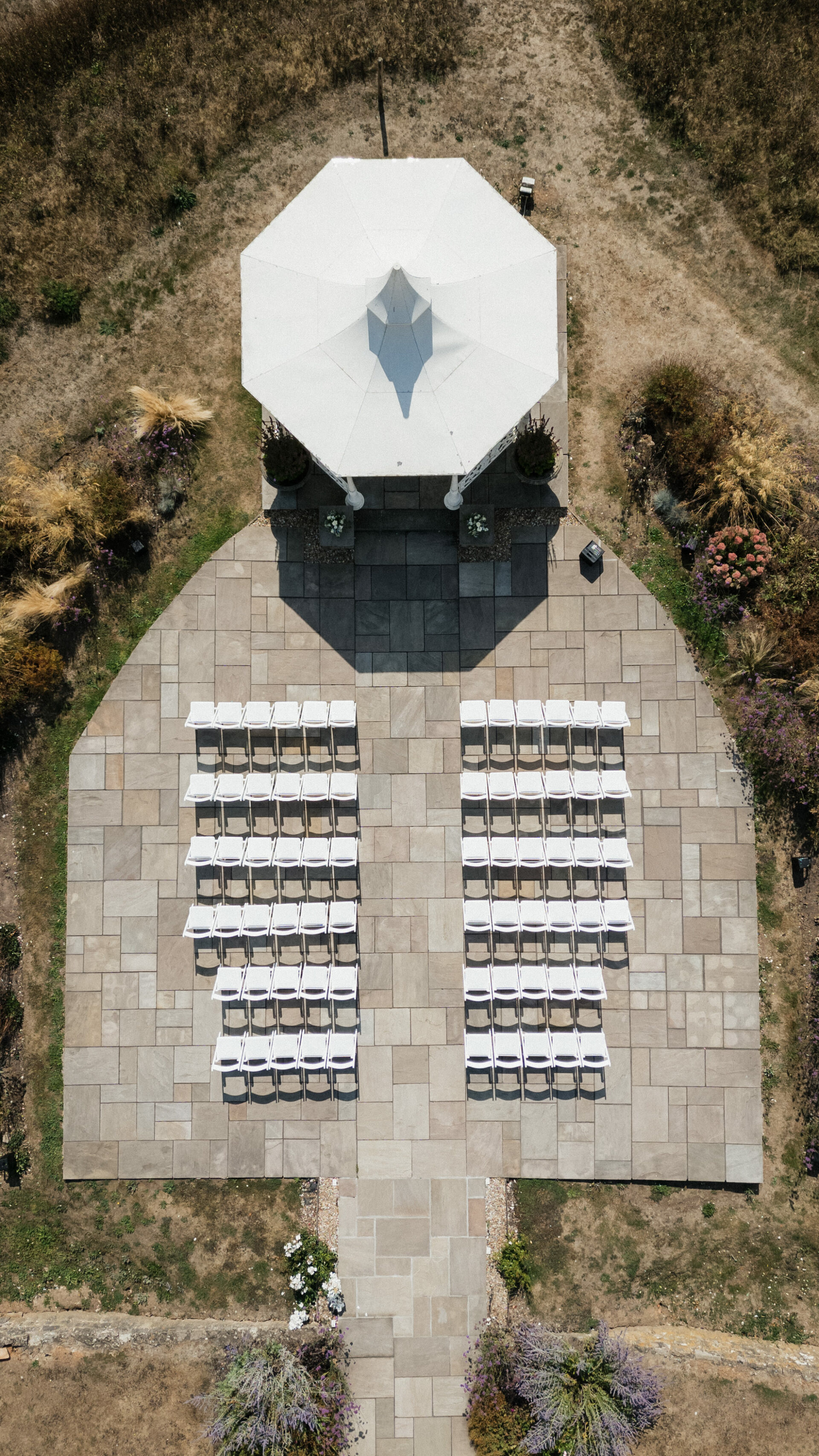View from above of a patio set up for an outdoor ceremony, with white chairs facing a gazebo, surrounded by dry grass and plants.