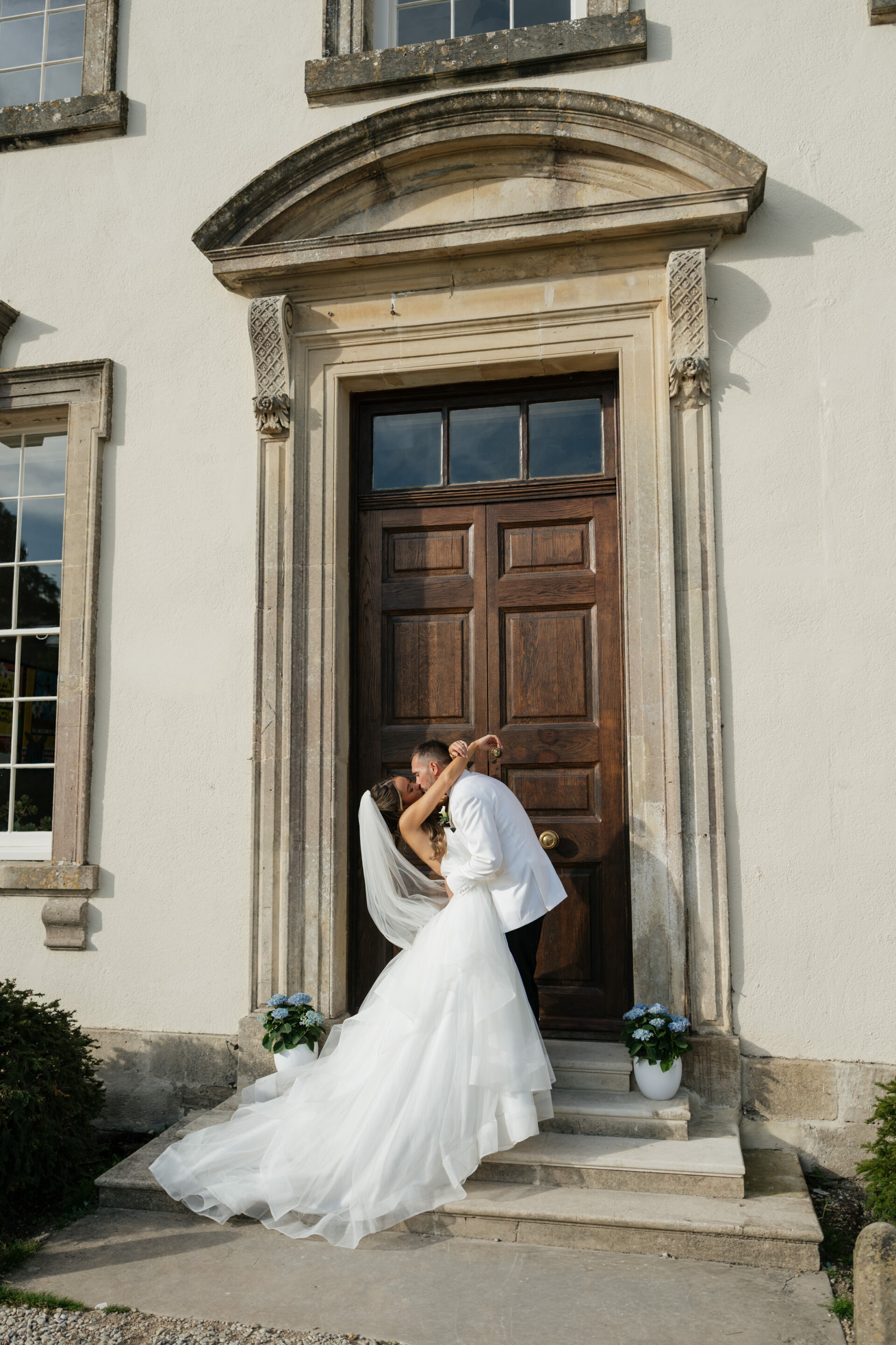 A bride and groom share a kiss on the steps outside a white building, with big wooden doors and potted plants by the entrance.