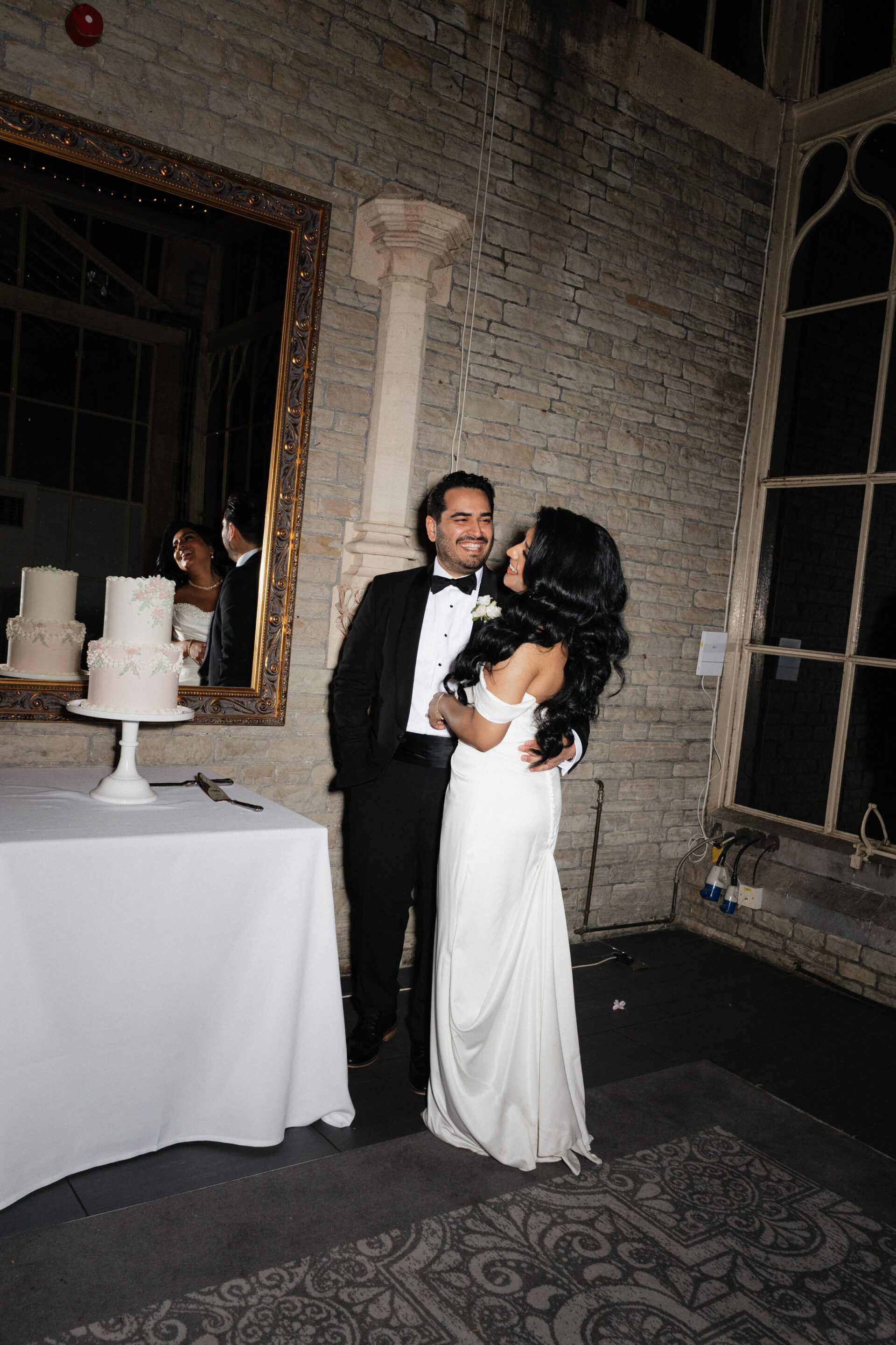 A dressed-up couple stands next to each other by a table with two white cakes, in a room with stone walls and big windows.