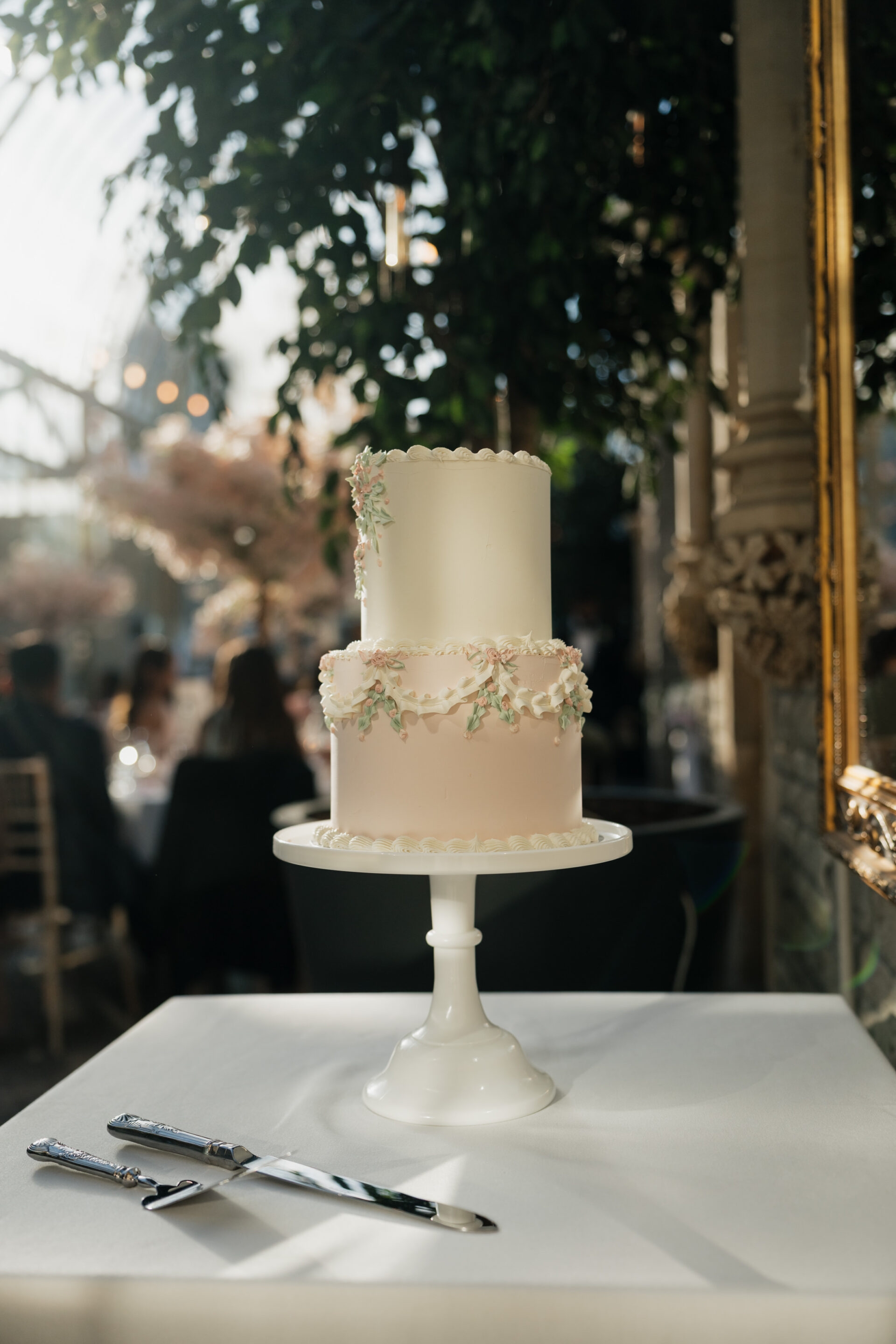 A white and pink two-layer cake with flowers sits on a white stand, ready to cut in a bright room with sunshine coming in.
