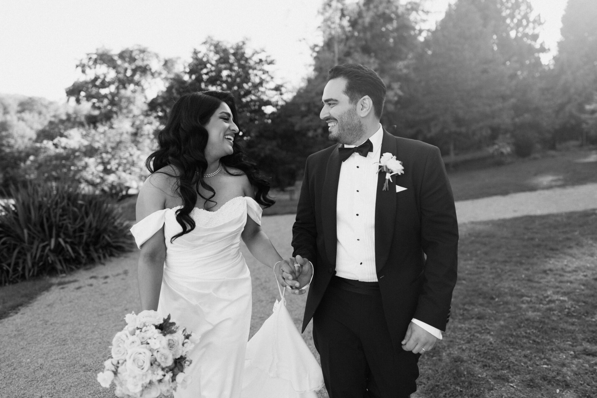A bride and groom dressed up nicely walk through a garden holding hands and smiling at each other.