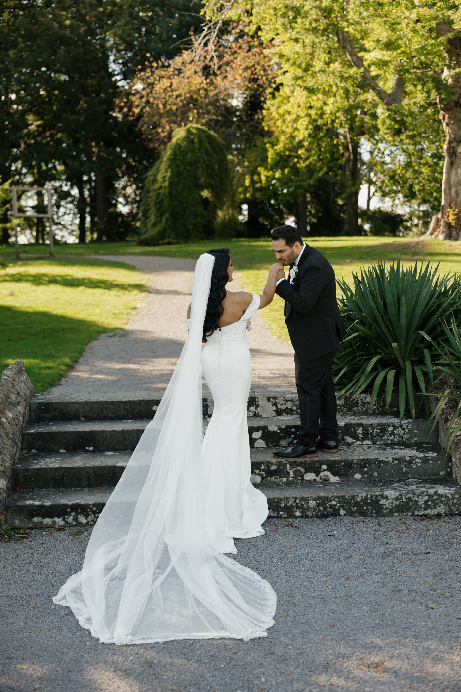 A groom in a black suit kisses his bride’s hand as she stands in a white dress and veil on stone steps with green plants around.