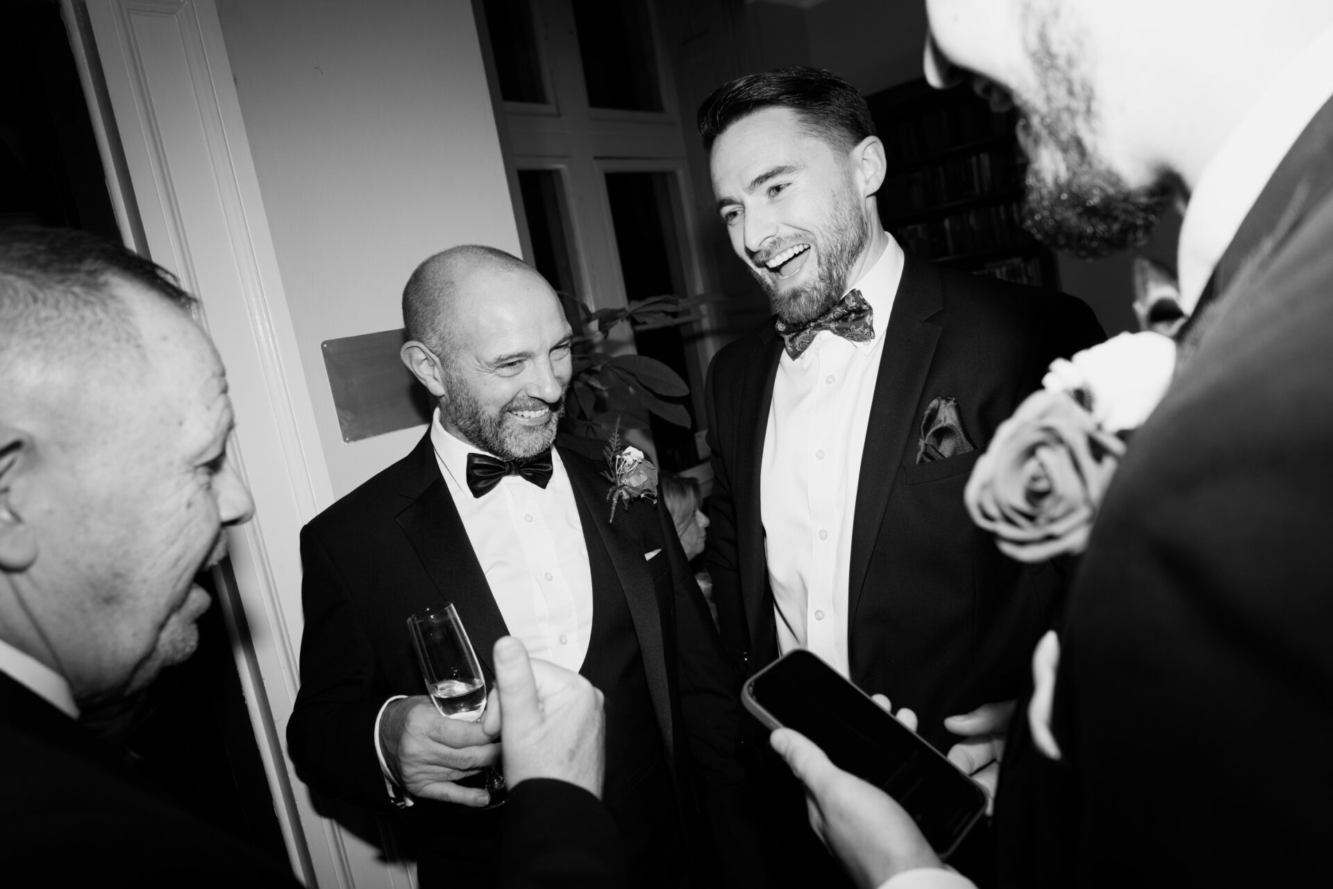 Four men dressed up, some with drinks in hand, are standing together inside, smiling and talking at what looks like a party.