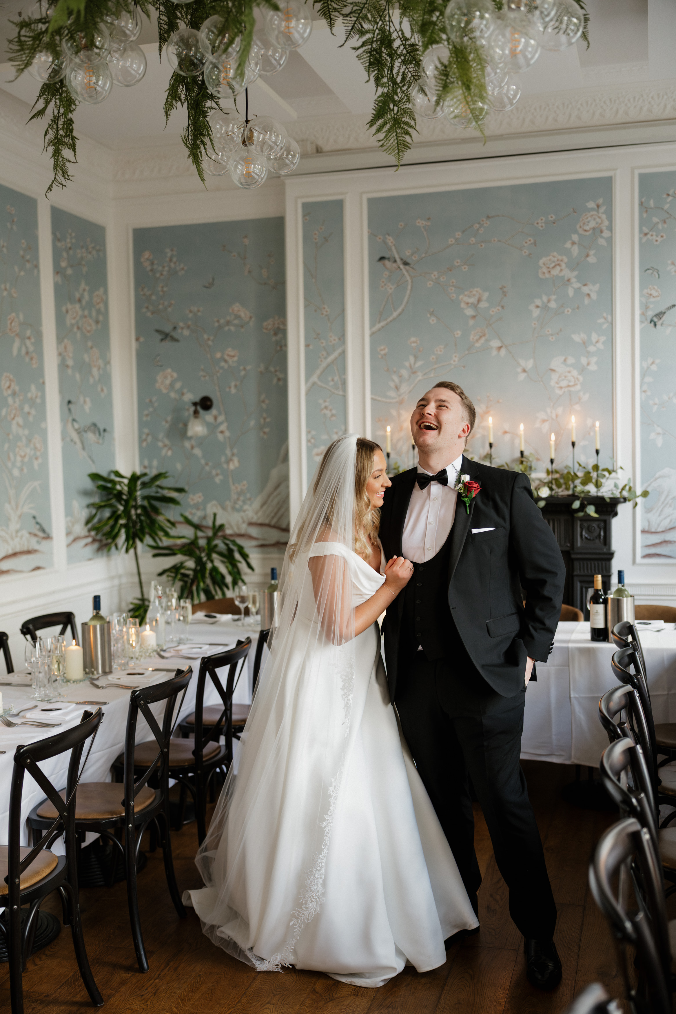 A bride and groom laugh together in a fancy dining room decorated with flowers and candles for a special occasion.