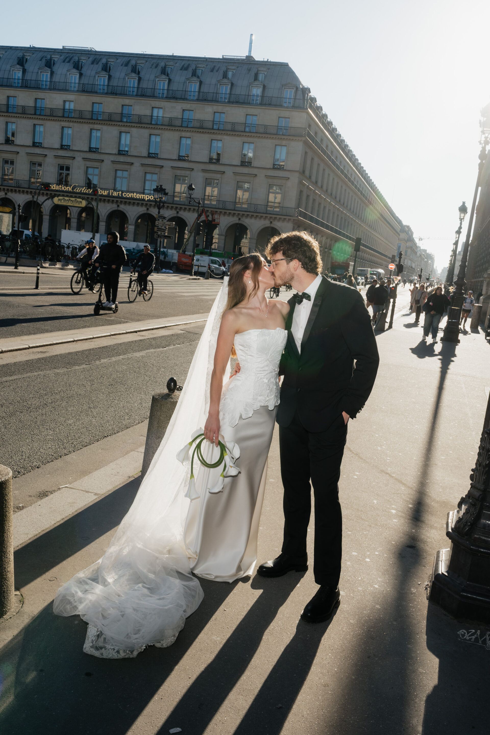 A bride and groom share a kiss on a sunny city sidewalk, holding flowers as people, cars, and bikes pass by in the background.