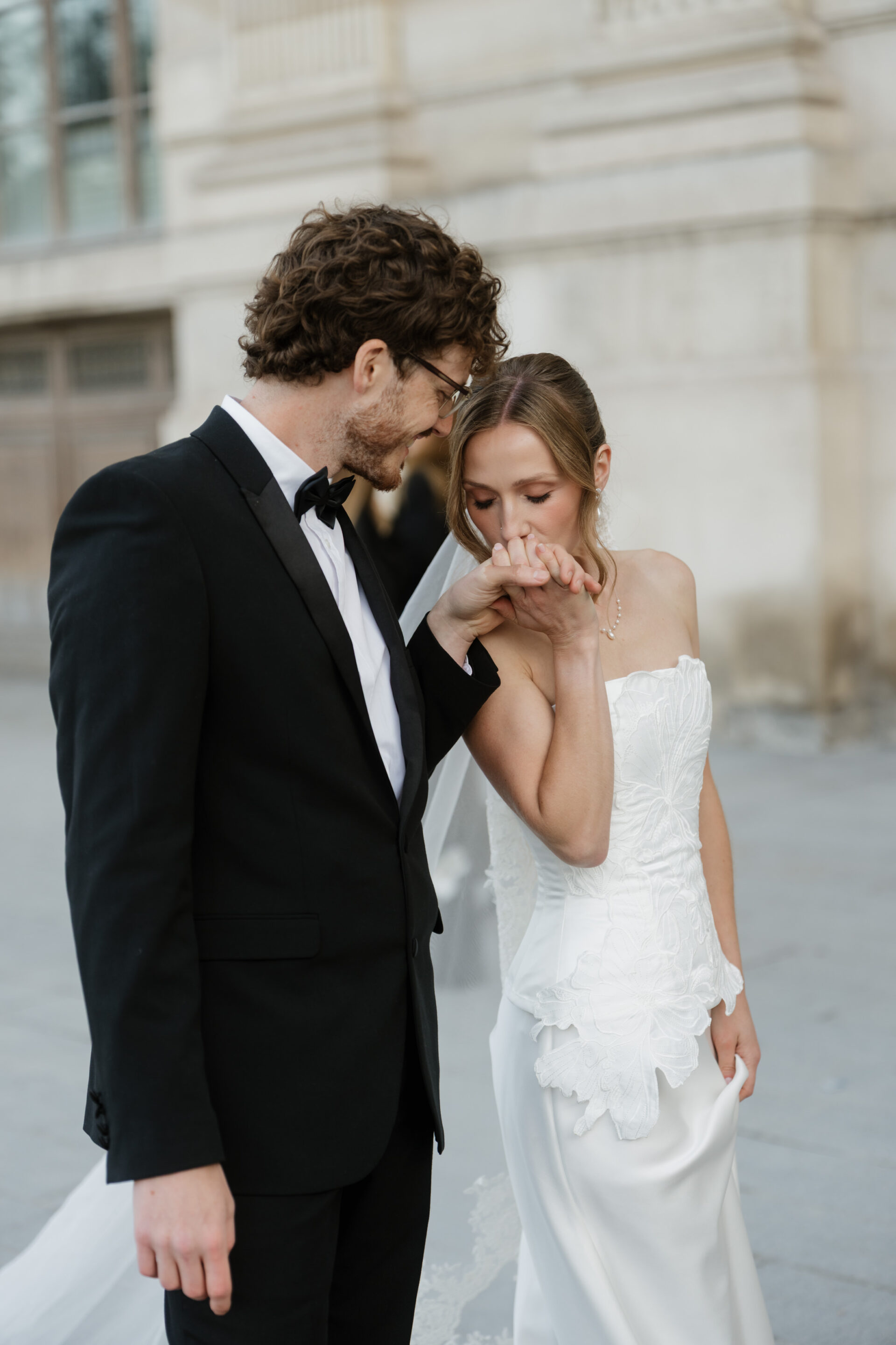 A man in a black suit kisses a woman’s hand. She’s wearing a white strapless wedding dress, and they’re outside by a stone building.