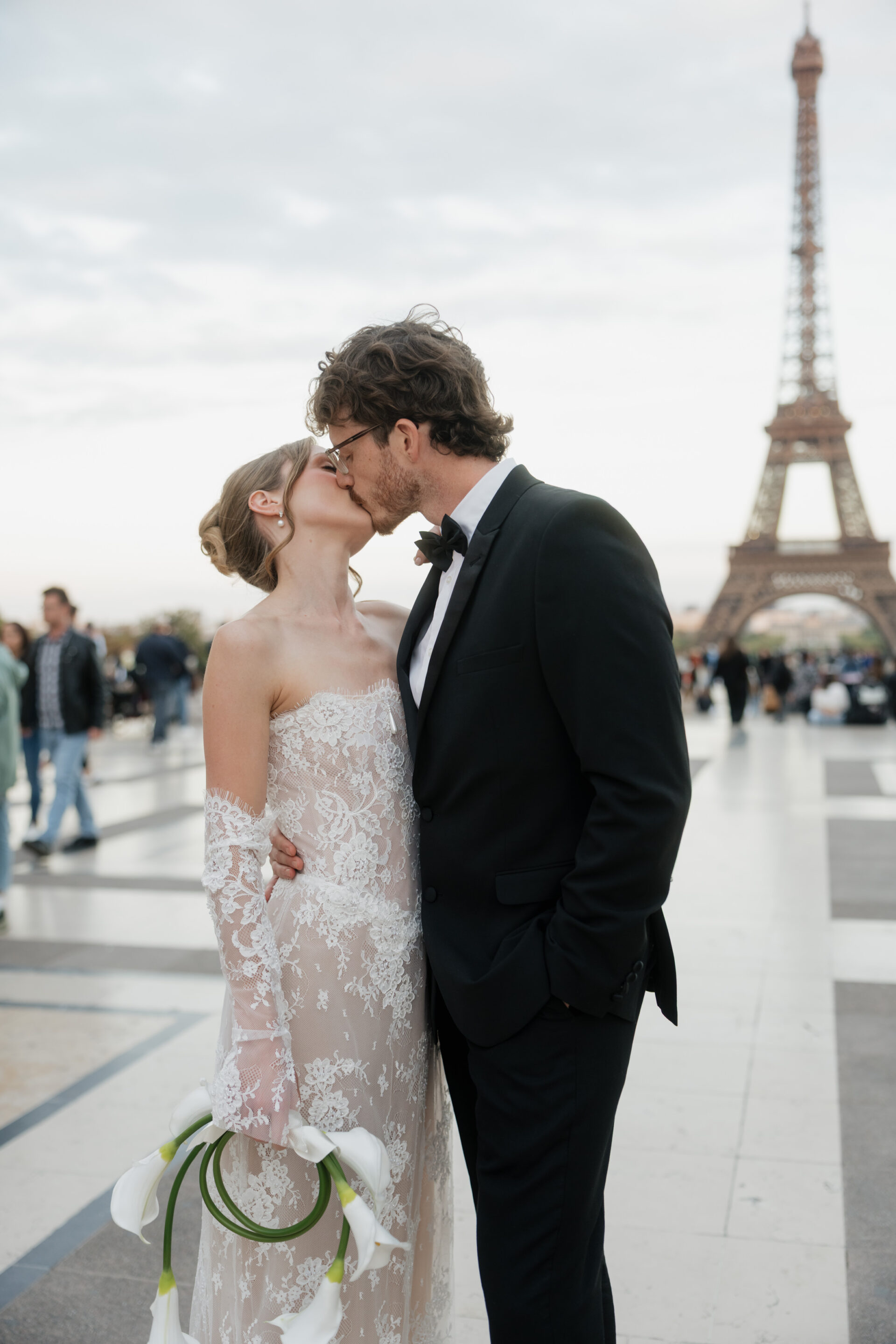 A bride and groom kiss by the Eiffel Tower, with others around them. The bride holds some white calla lilies.