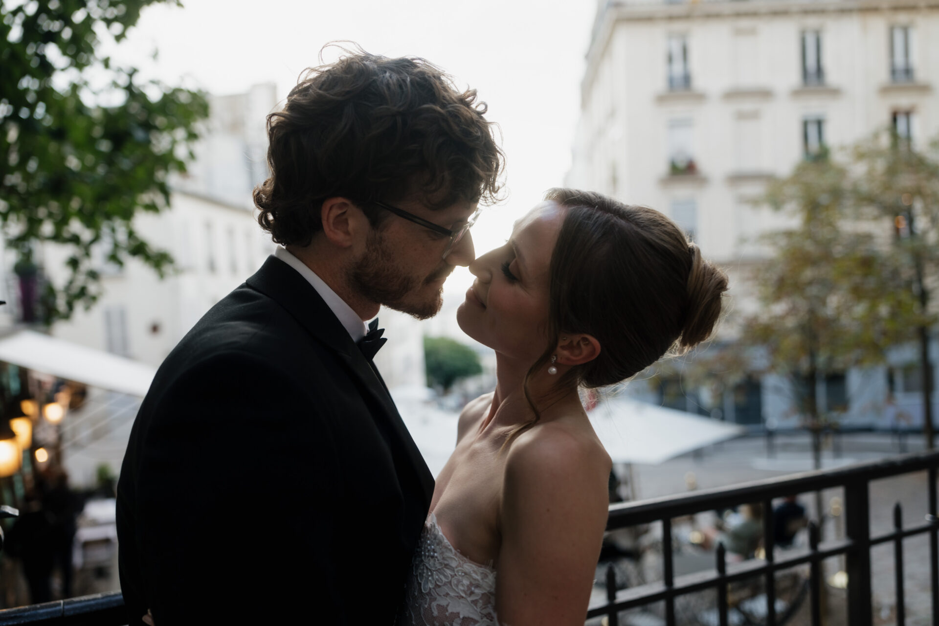 A bride and groom are standing outside, gazing into each other's eyes, with some trees and city buildings behind them.