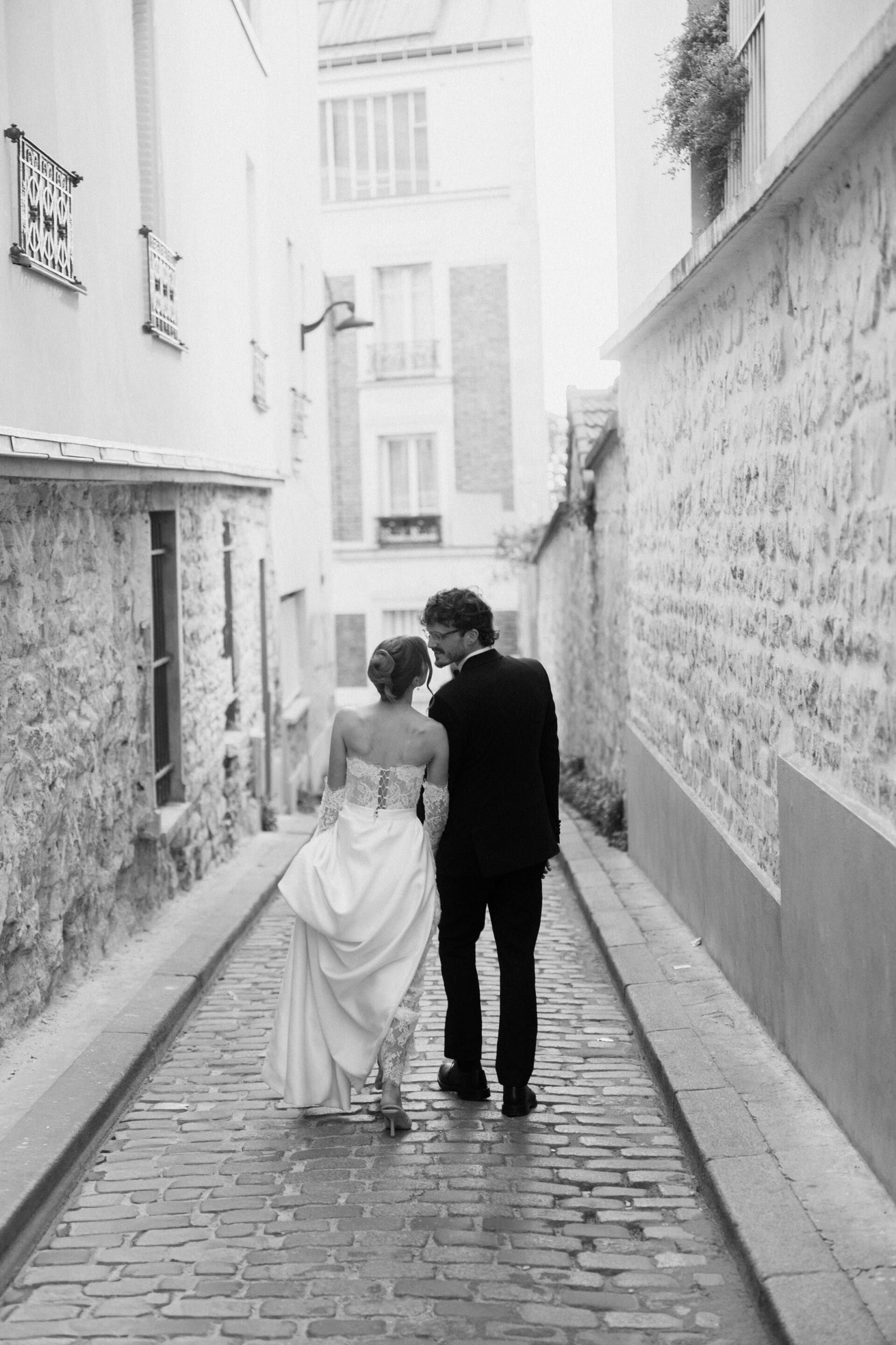 A bride and groom walk together, arm in arm, down a narrow old street with stone walls, seen from behind in black and white.