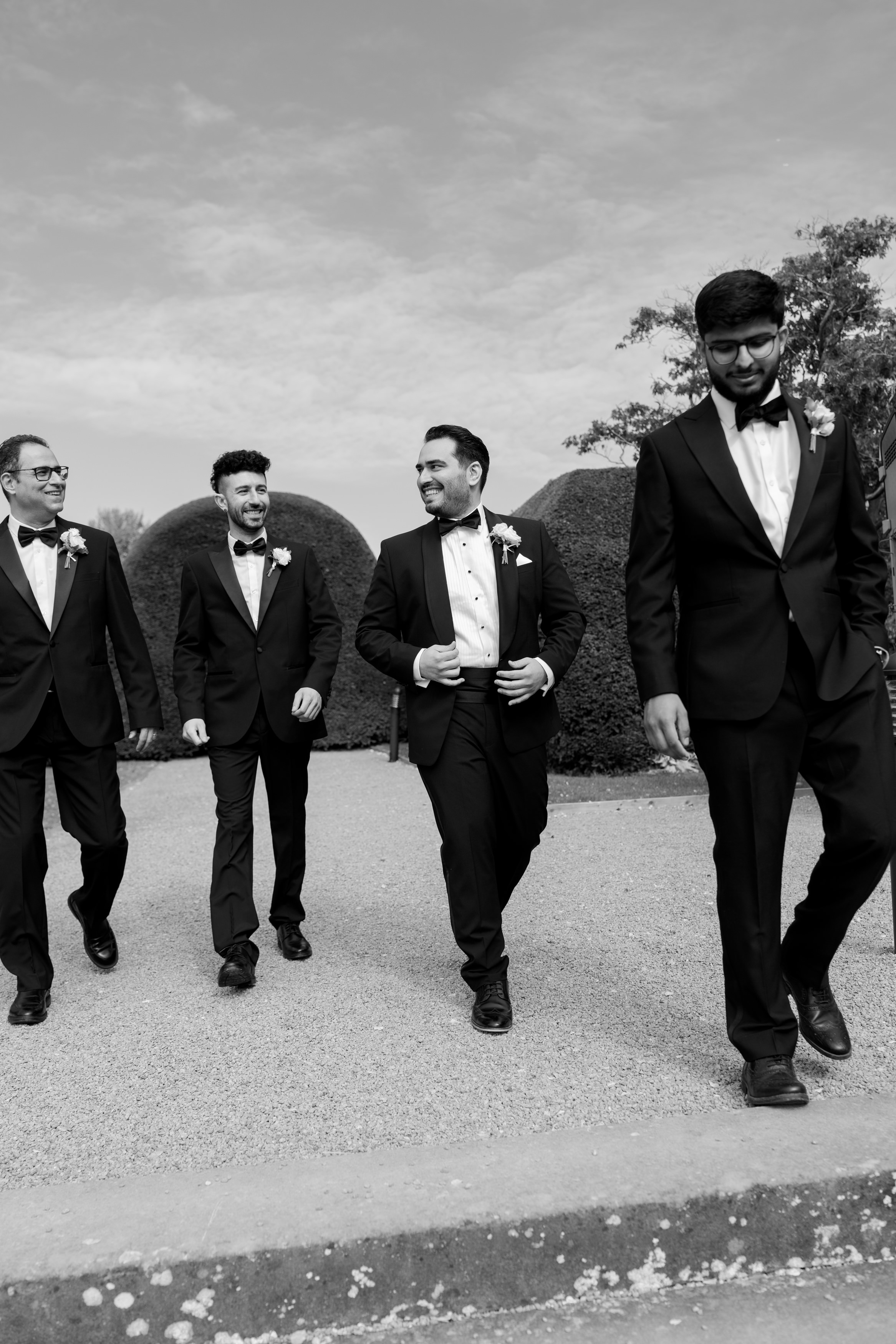 Four men in suits and bow ties are walking outside on a gravel path, chatting and smiling, with bushes and clear sky behind them.