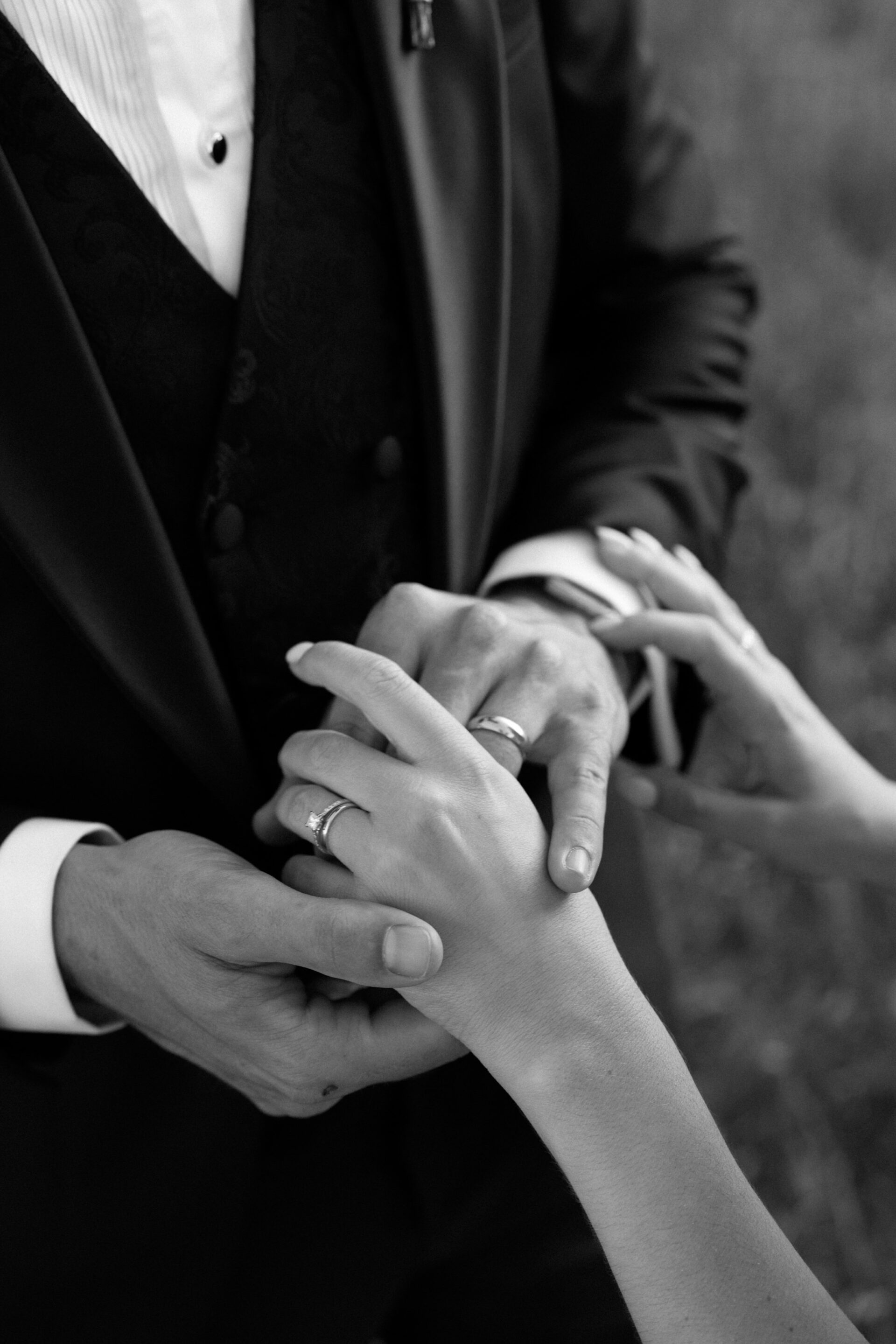 Two people are holding hands, both wearing rings. One is wearing a suit. The photo is in black and white.