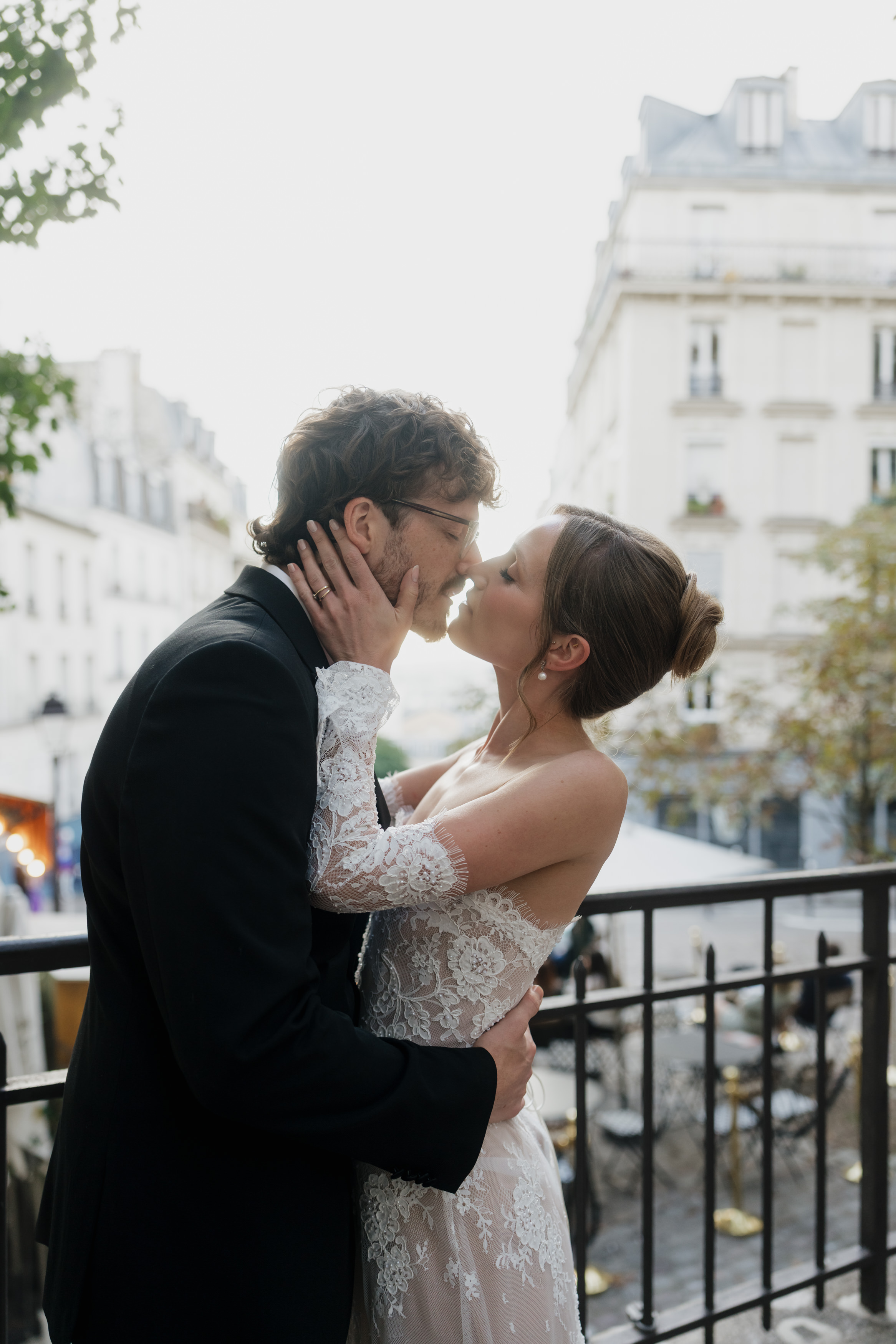 A bride and groom share a kiss outside, standing close on a balcony with European-looking buildings behind them.