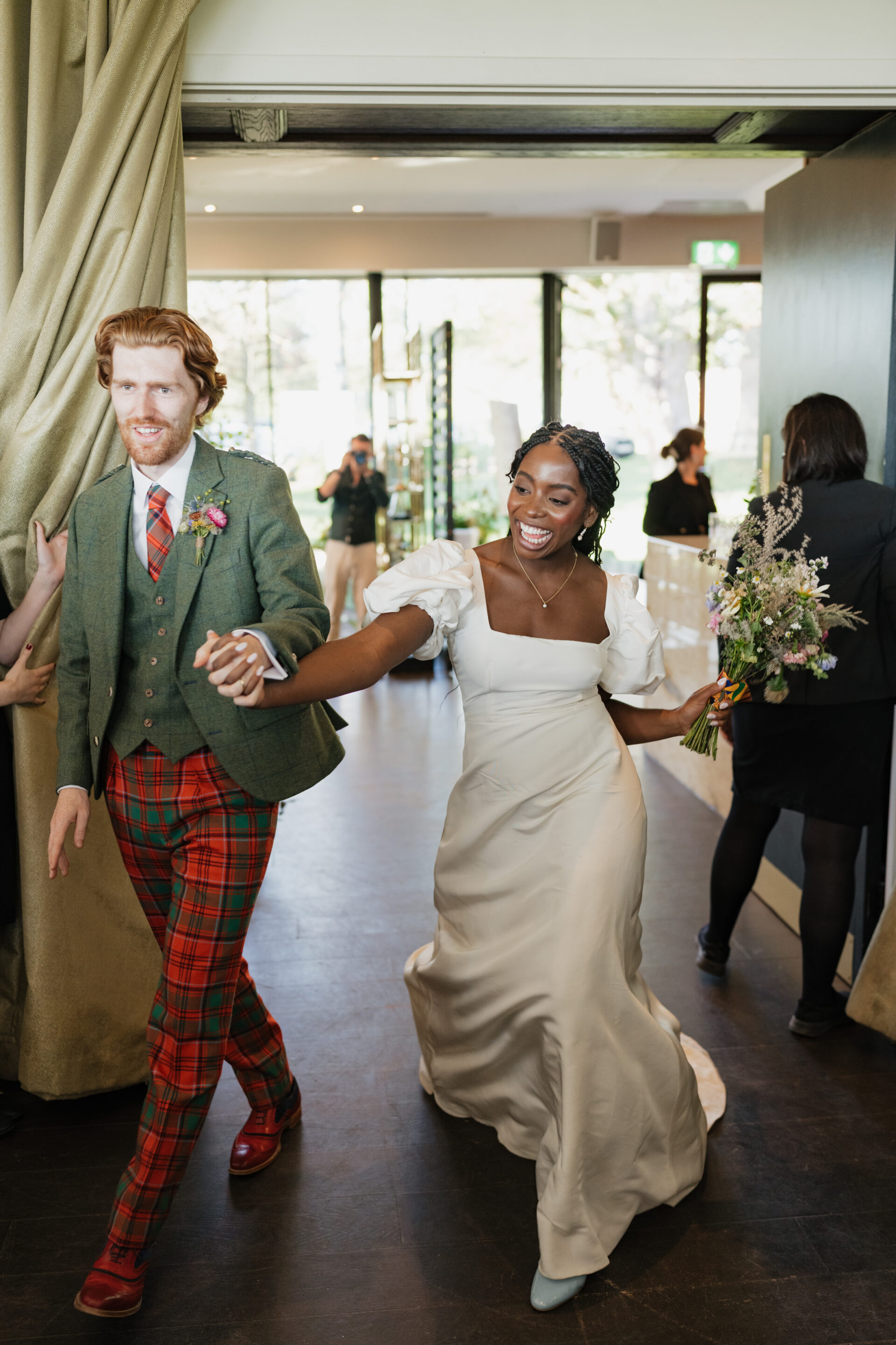 A newly married couple walks into the room, holding hands and smiling. The groom is dressed in a green jacket and red plaid pants, while the bride is wearing a white dress and carrying a bouquet of flowers.