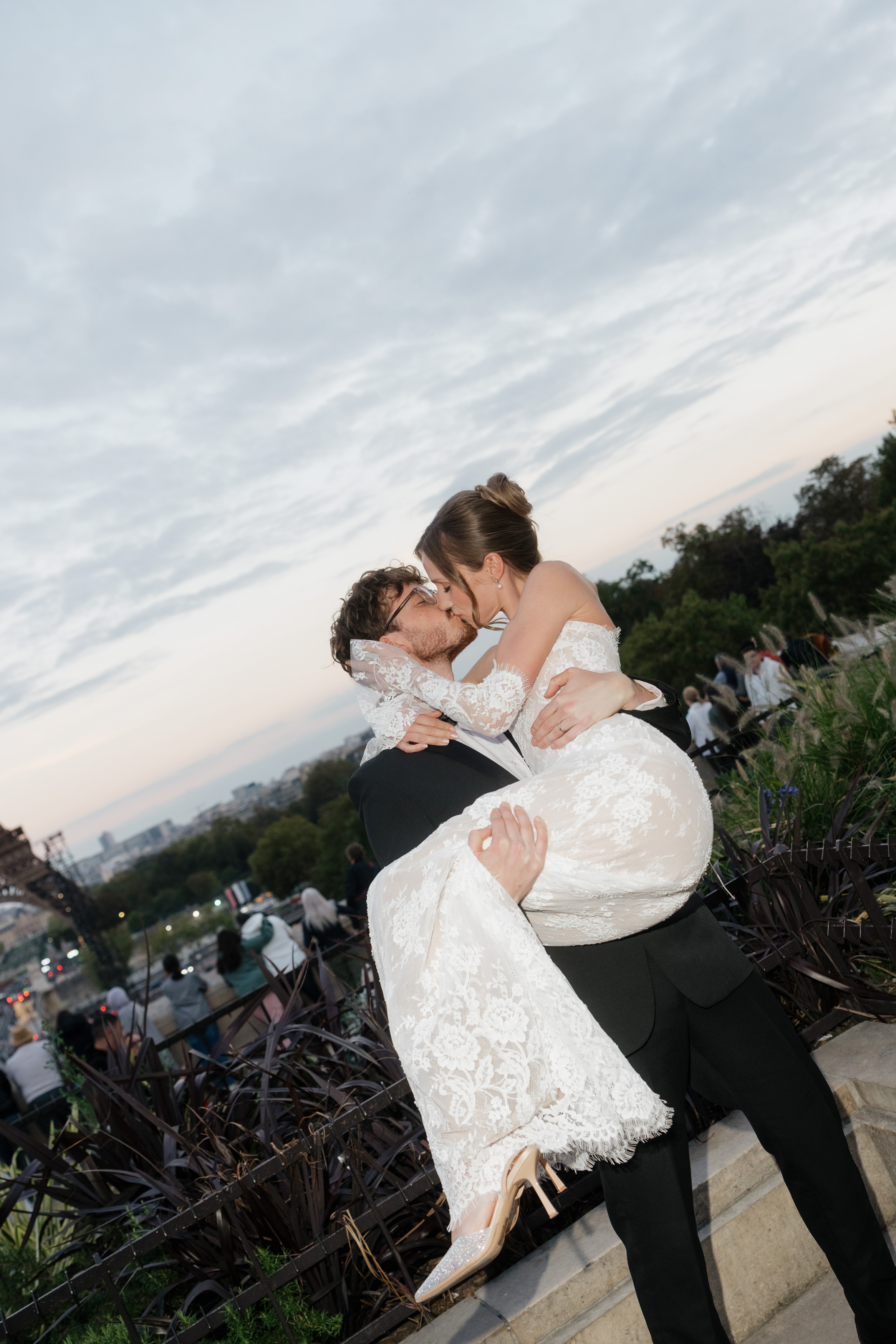 A groom in a black suit lifts up and kisses his bride, who’s wearing a white lace dress, outside with trees, people, and city buildings nearby.