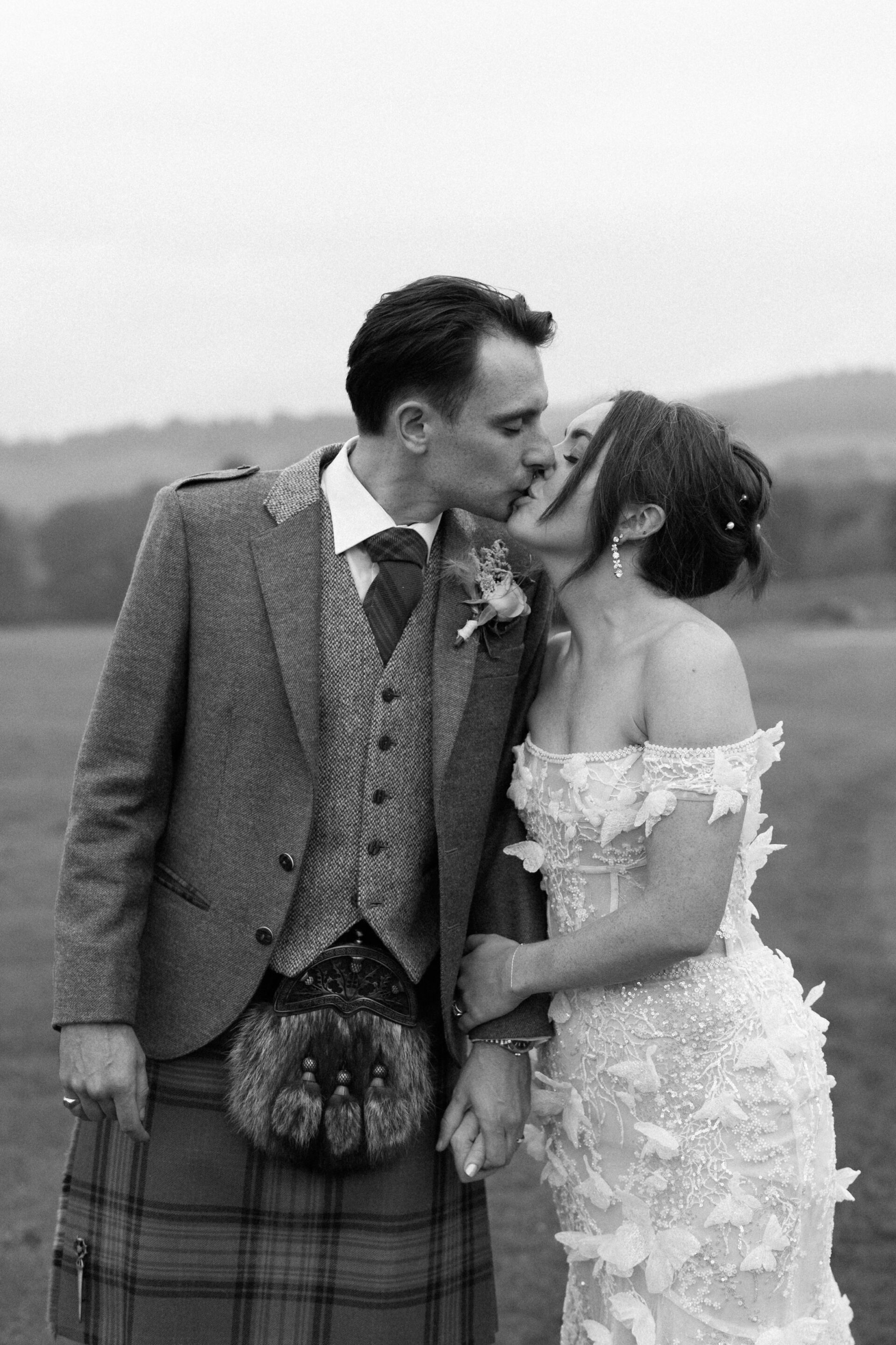 A bride and groom, dressed for their wedding, kiss outside. The groom is wearing a kilt and jacket, while the bride has on an off-the-shoulder lace dress.