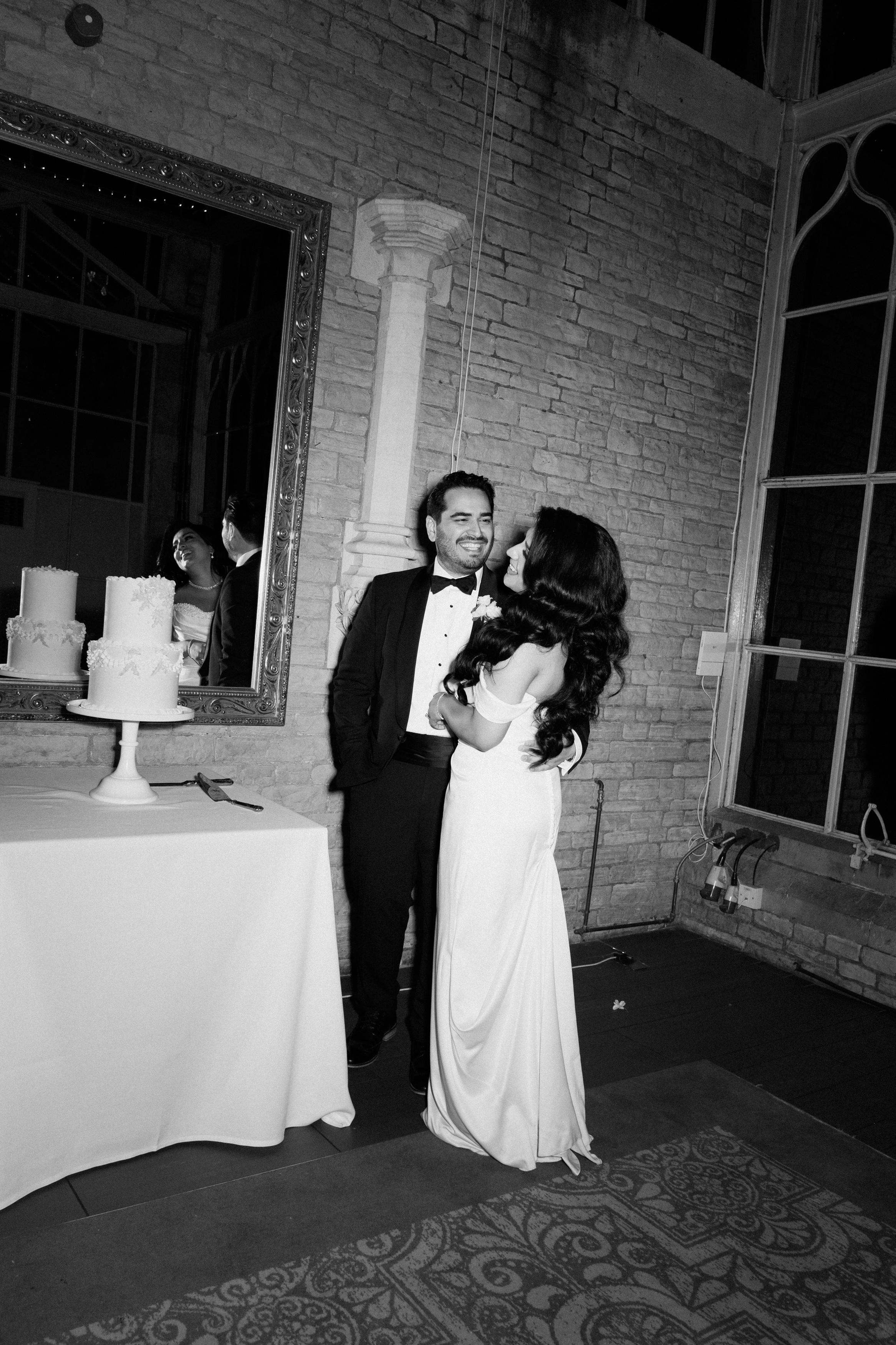A dressed-up couple stands by a table with two cakes, in front of a big mirror and brick wall at an indoor gathering.