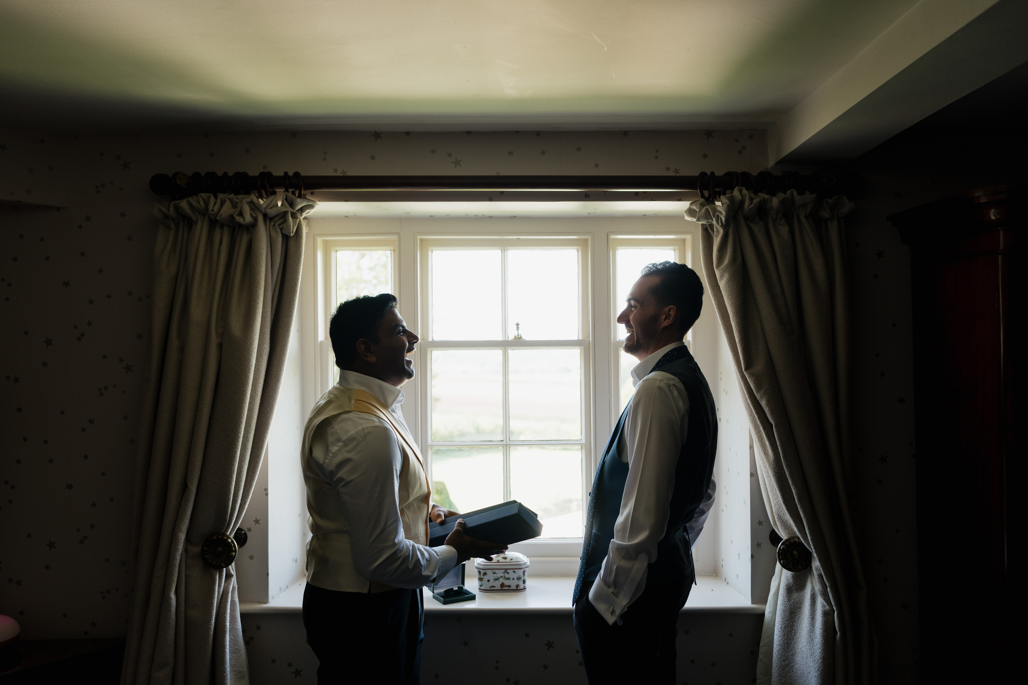 Two men dressed up nicely stand by a bright window, smiling at each other. One holds a book, and cream curtains hang around the window.