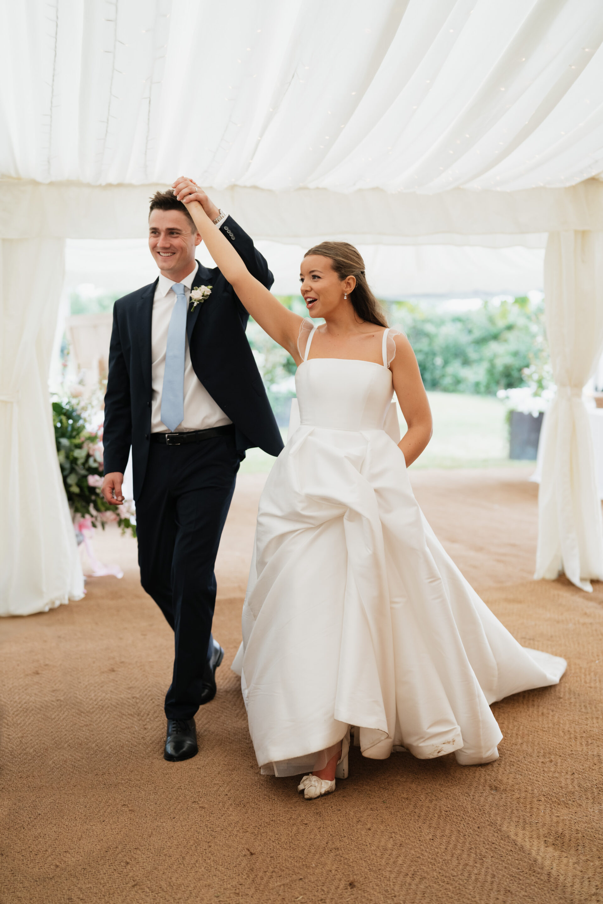 A bride and groom walk into their wedding under a white tent, holding hands. The bride is smiling and raising her arm.
