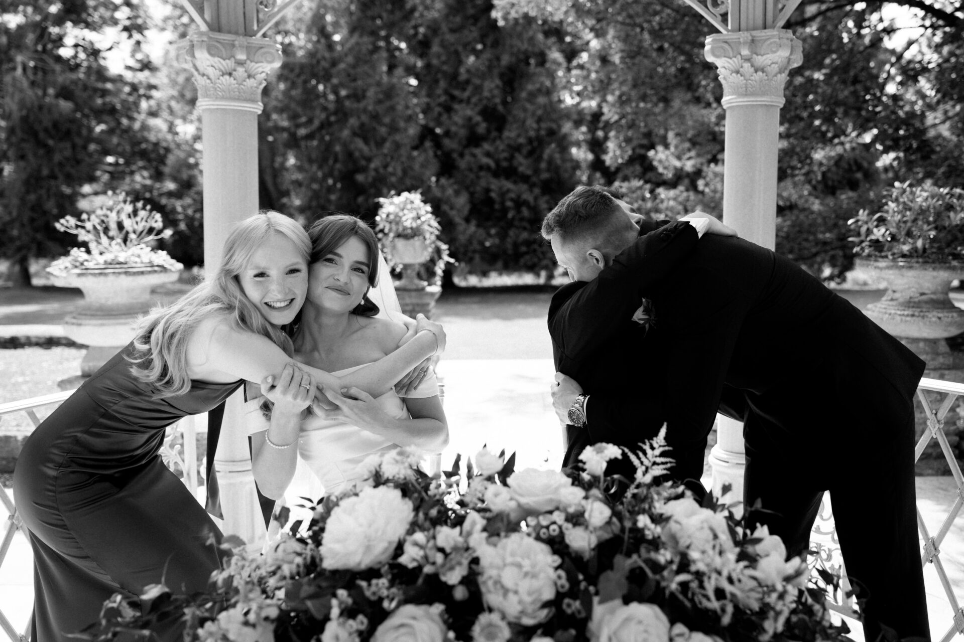 At a wedding, two women are sitting and hugging on the left, while two men hug on the right. Flowers sit in front of them.
