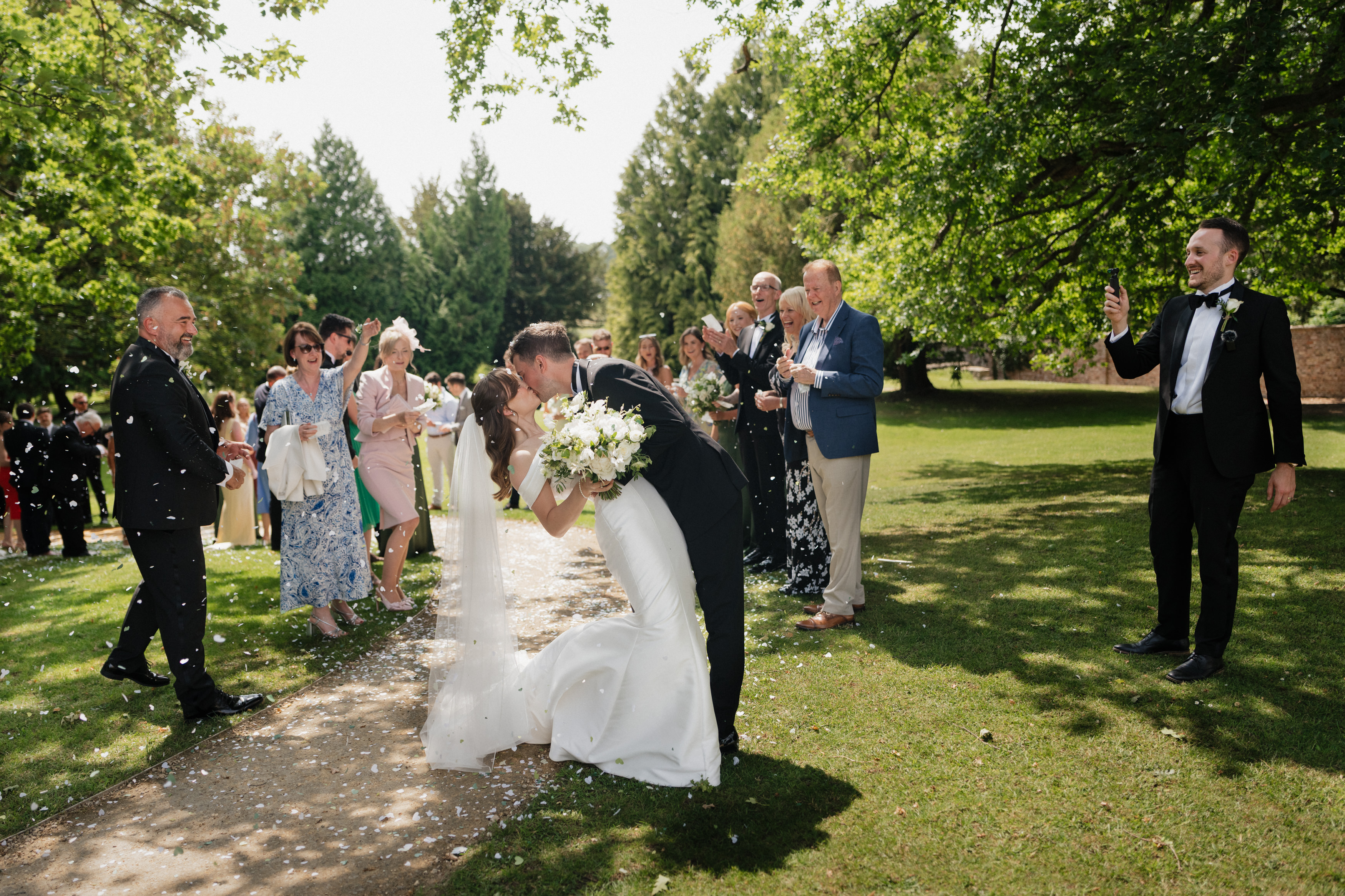 A bride and groom kiss outside on a path while their guests stand around, some clapping, others snapping pictures, with green trees all around.